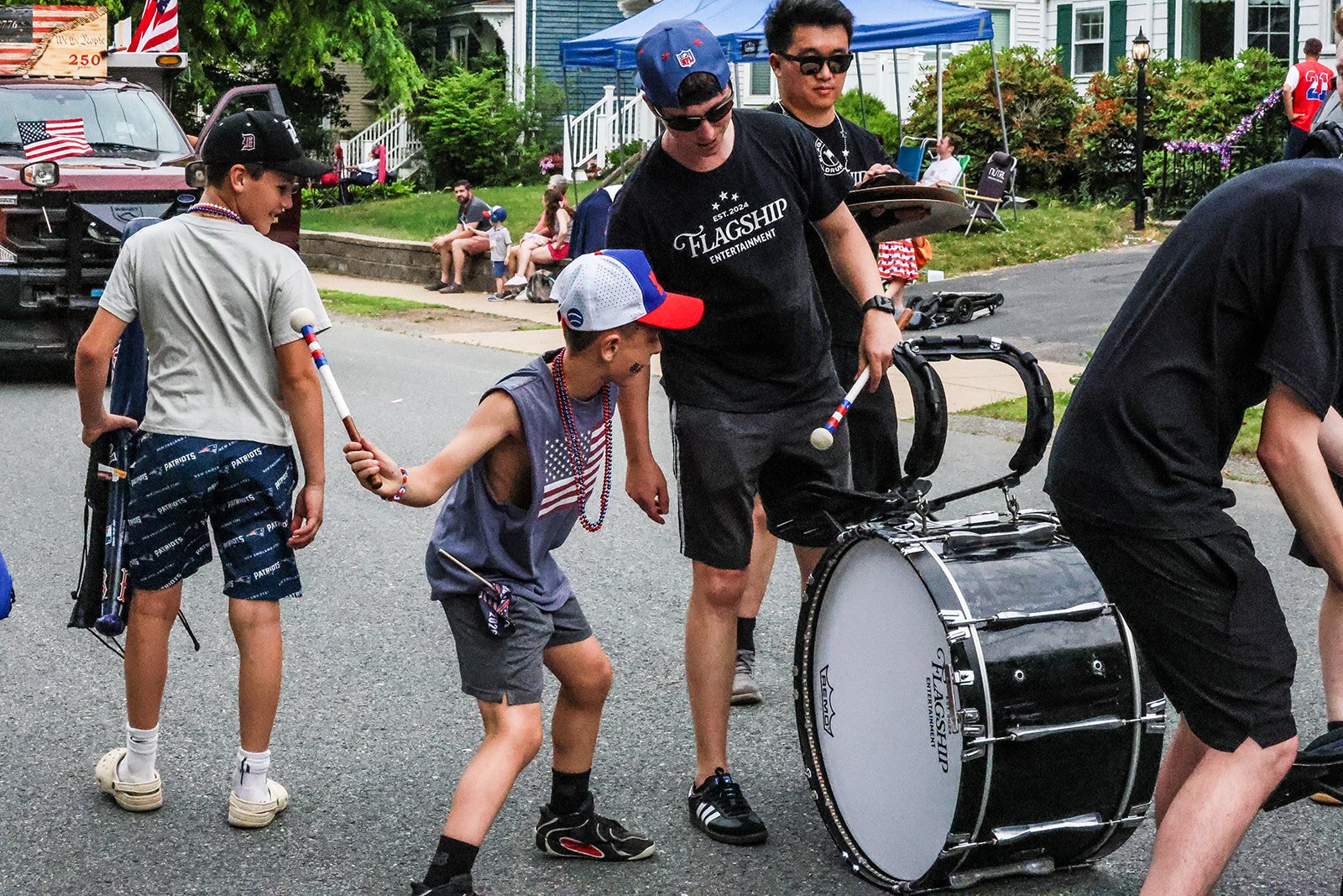 Children and adults participating in a parade or celebration, playing drums and carrying flags, with decorated floats and spectators in background.