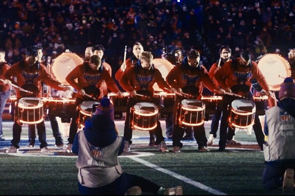 A group of drummers performing on a football field during a night game, with an audience in the background. They are wearing red jackets and playing illuminated drums, with media personnel in the foreground.