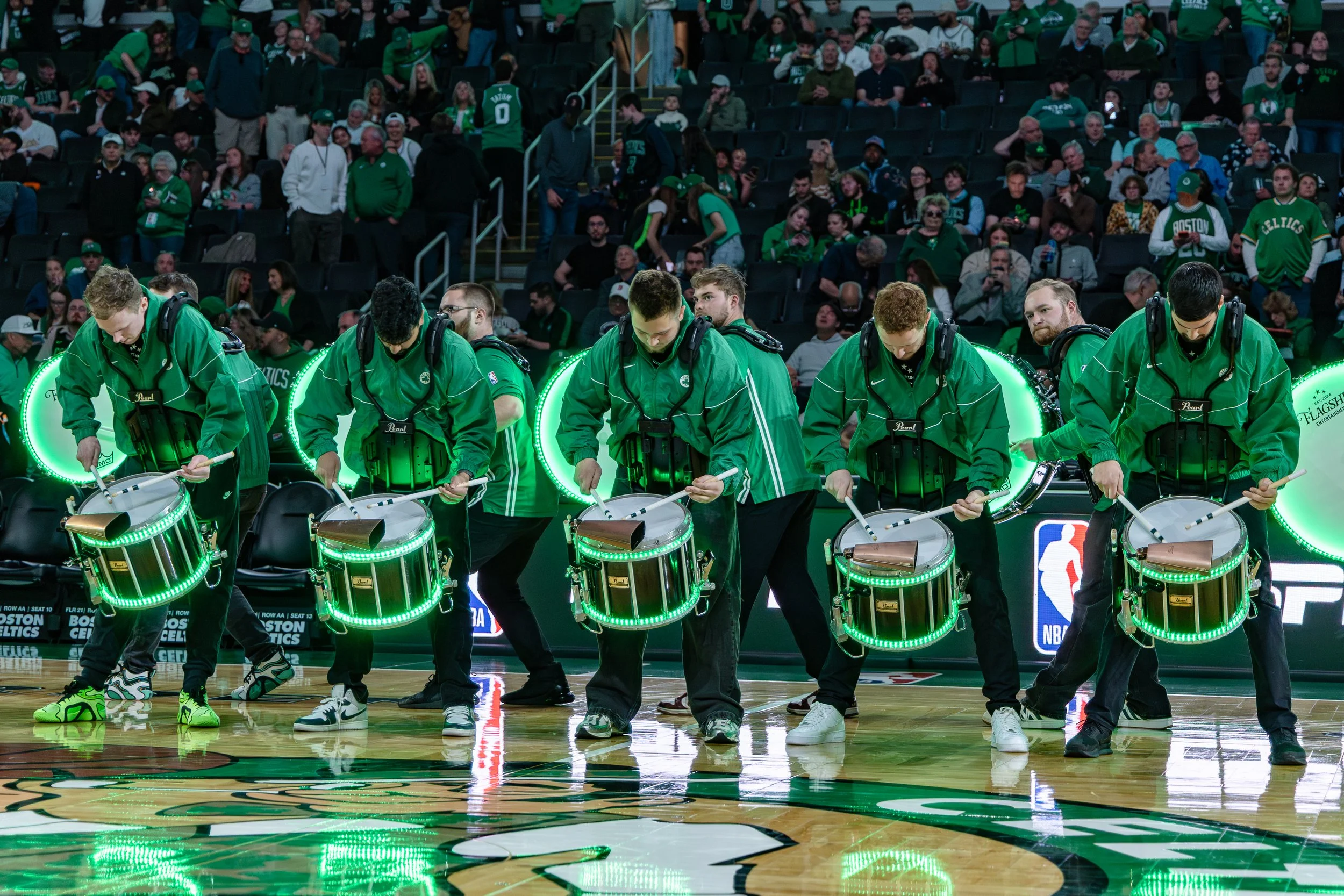 Celtics Basketball arena with a group of drummers in green jackets performing on the court, wearing headphones, surrounded by an audience mostly dressed in green, with some standing and others seated, watching the performance.