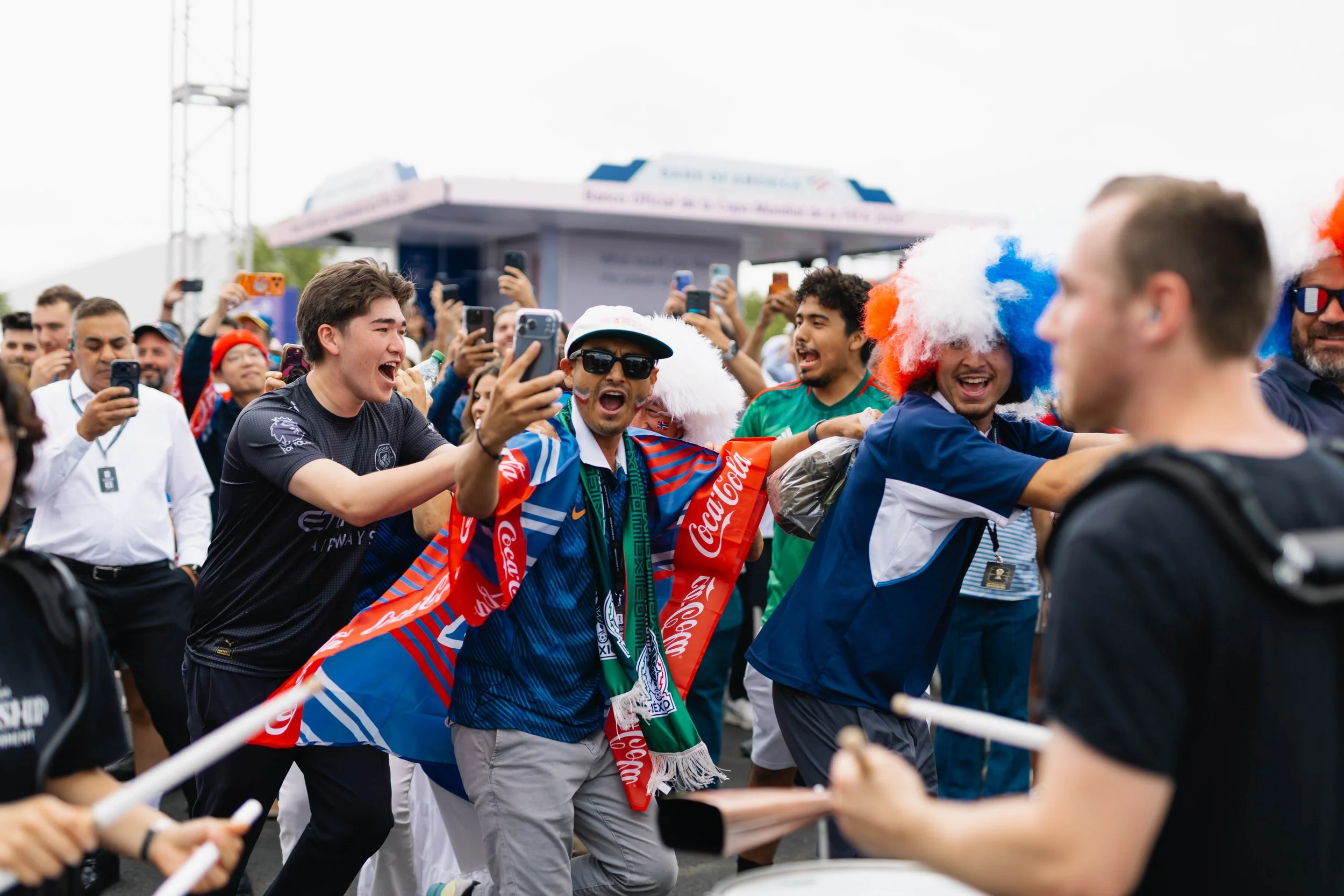 Crowd of fans celebrating and taking selfies at a sporting event, with some wearing Mexican flags and wigs in the colors of the Mexican flag.
