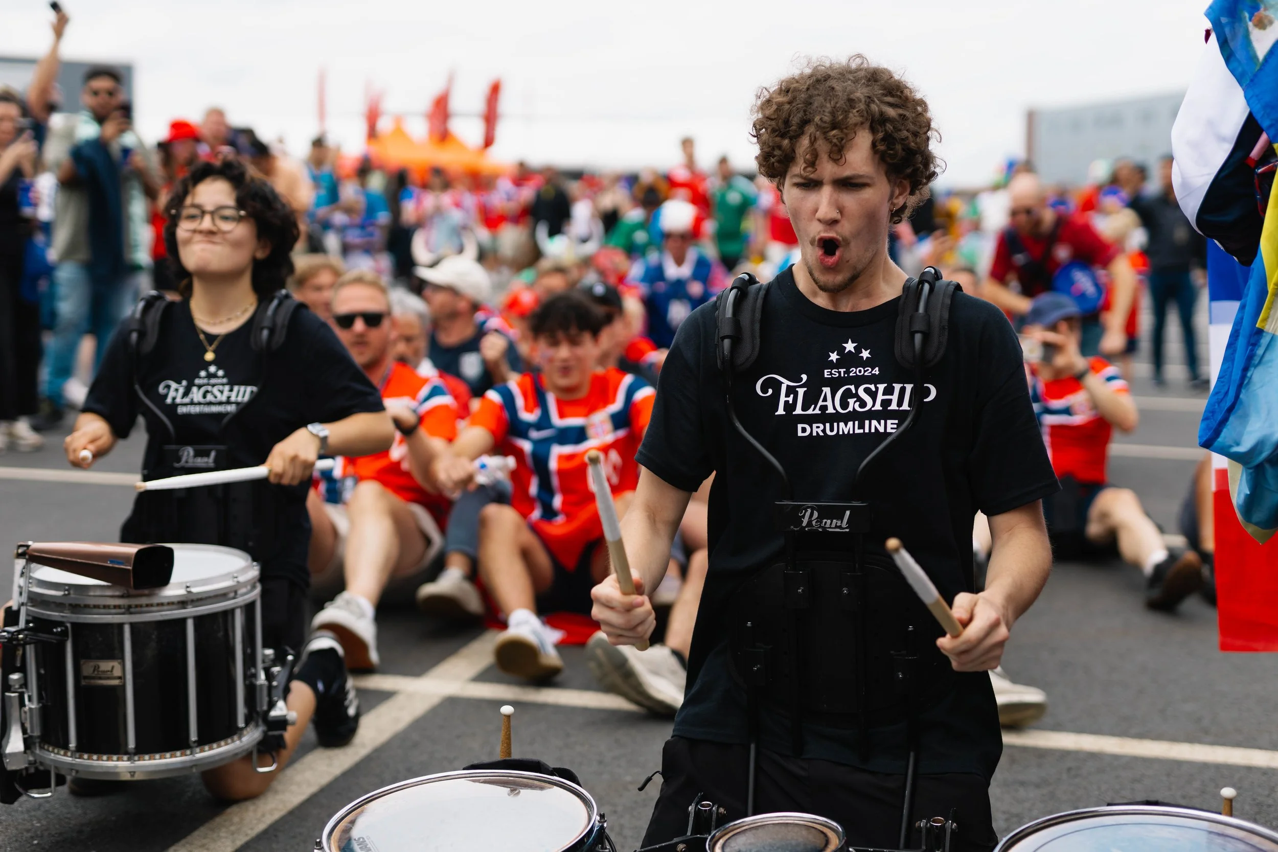 A young man with curly hair playing drums at an FIFA WORLD CUP event, surrounded by a crowd of people, some wearing red, blue, green, and black shirts, and a colorful amusement ride in the background.