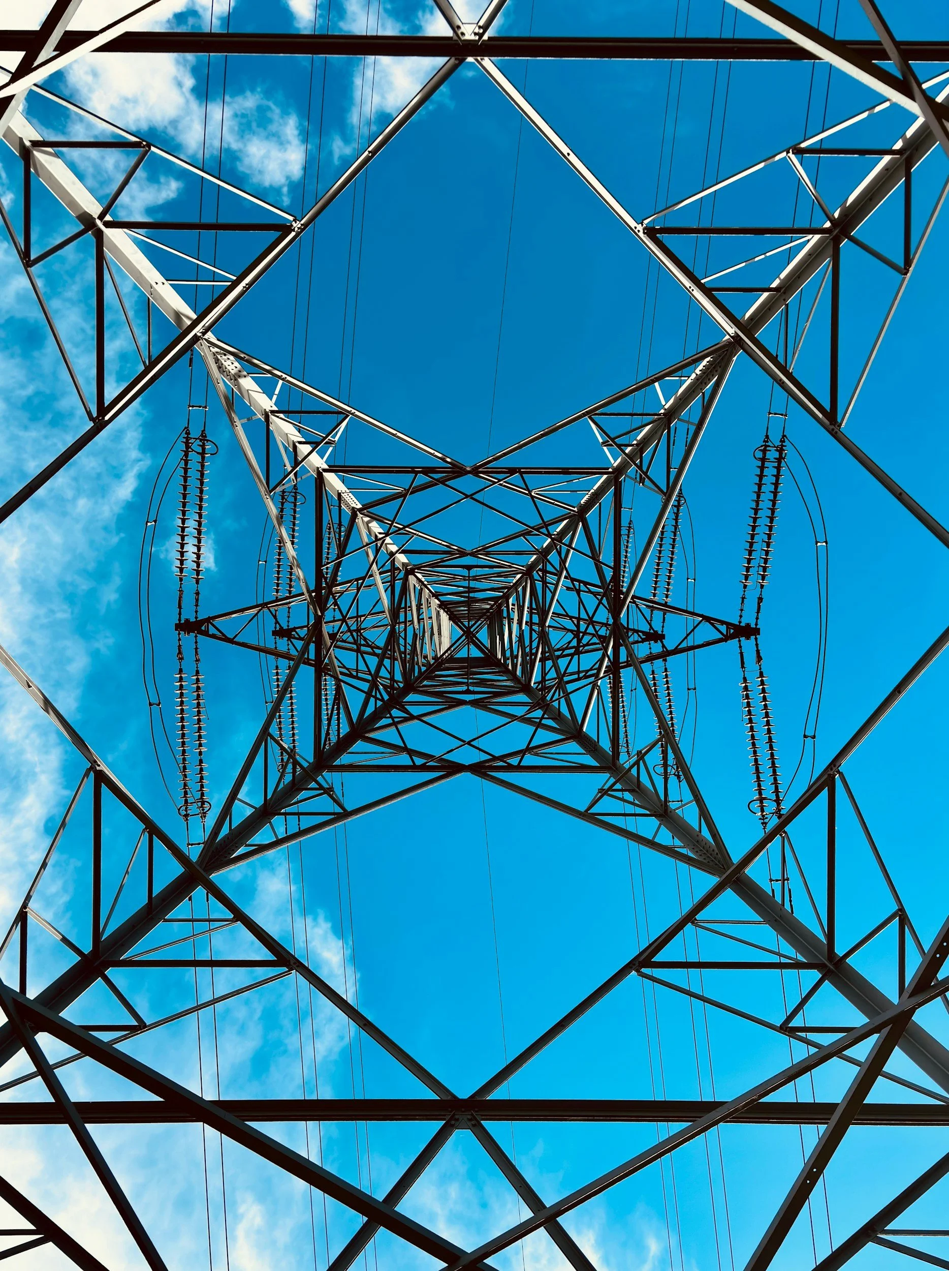 Looking up at a metal electrical transmission tower against a blue sky with scattered clouds.