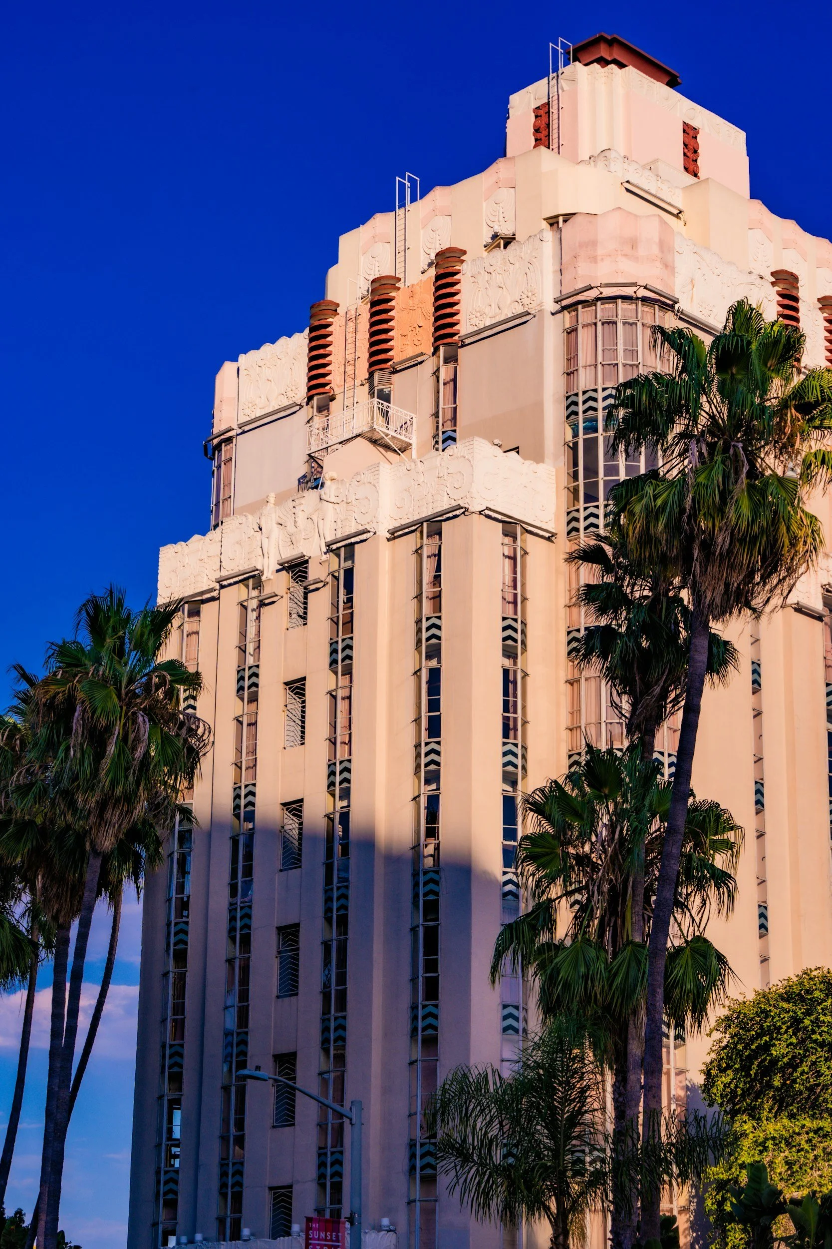 A tall, pink art deco building with intricate designs and multiple vertical windows, set against a vibrant blue sky with green palm trees in the foreground.