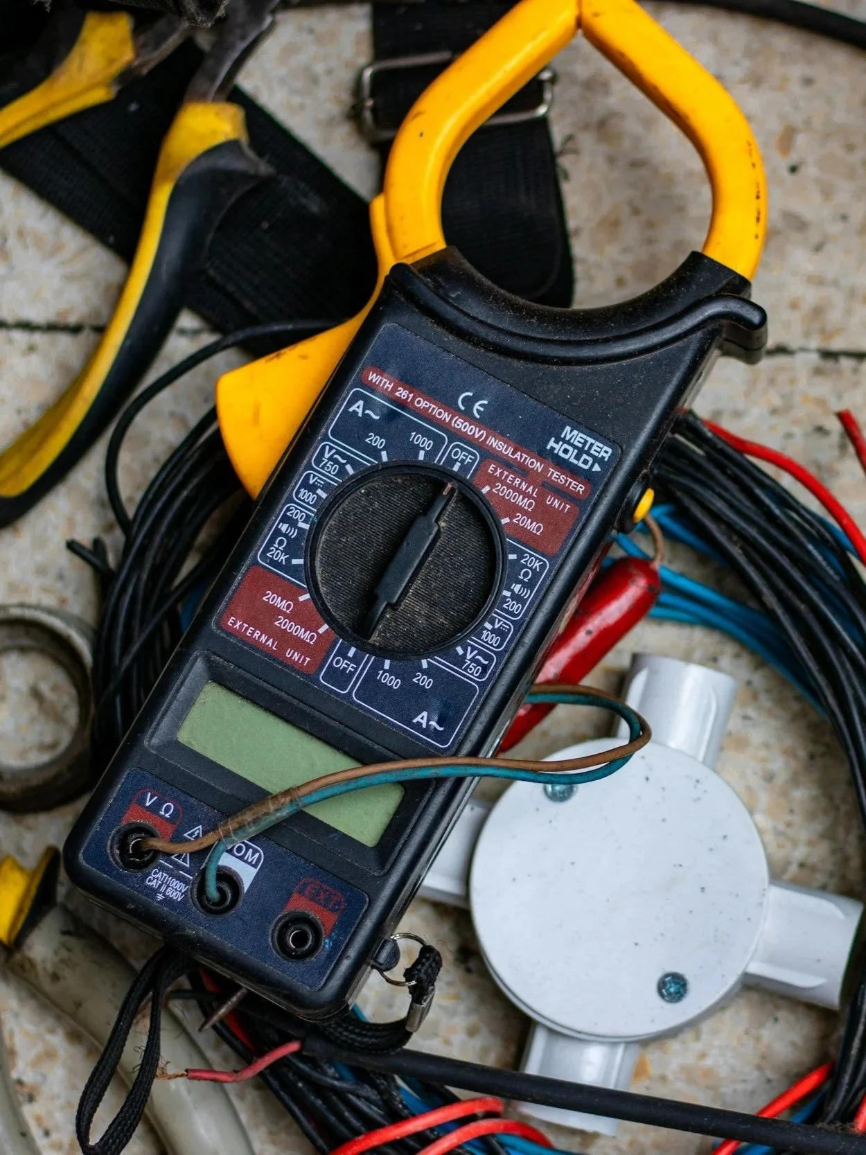 Digital multimeter with yellow and black probes, surrounded by electrical wires and a white electrical outlet, on a tiled floor.