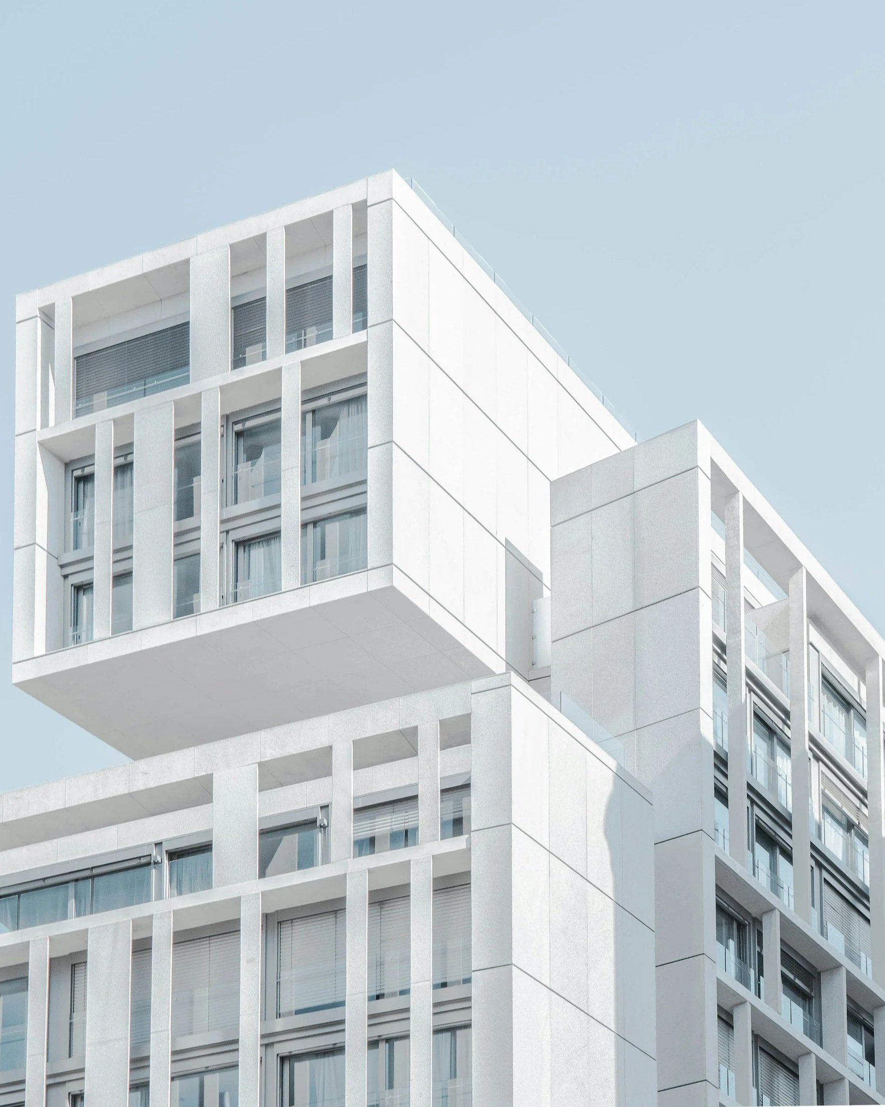 Modern white multi-story building with large glass windows and decorative vertical beams, under a clear blue sky.