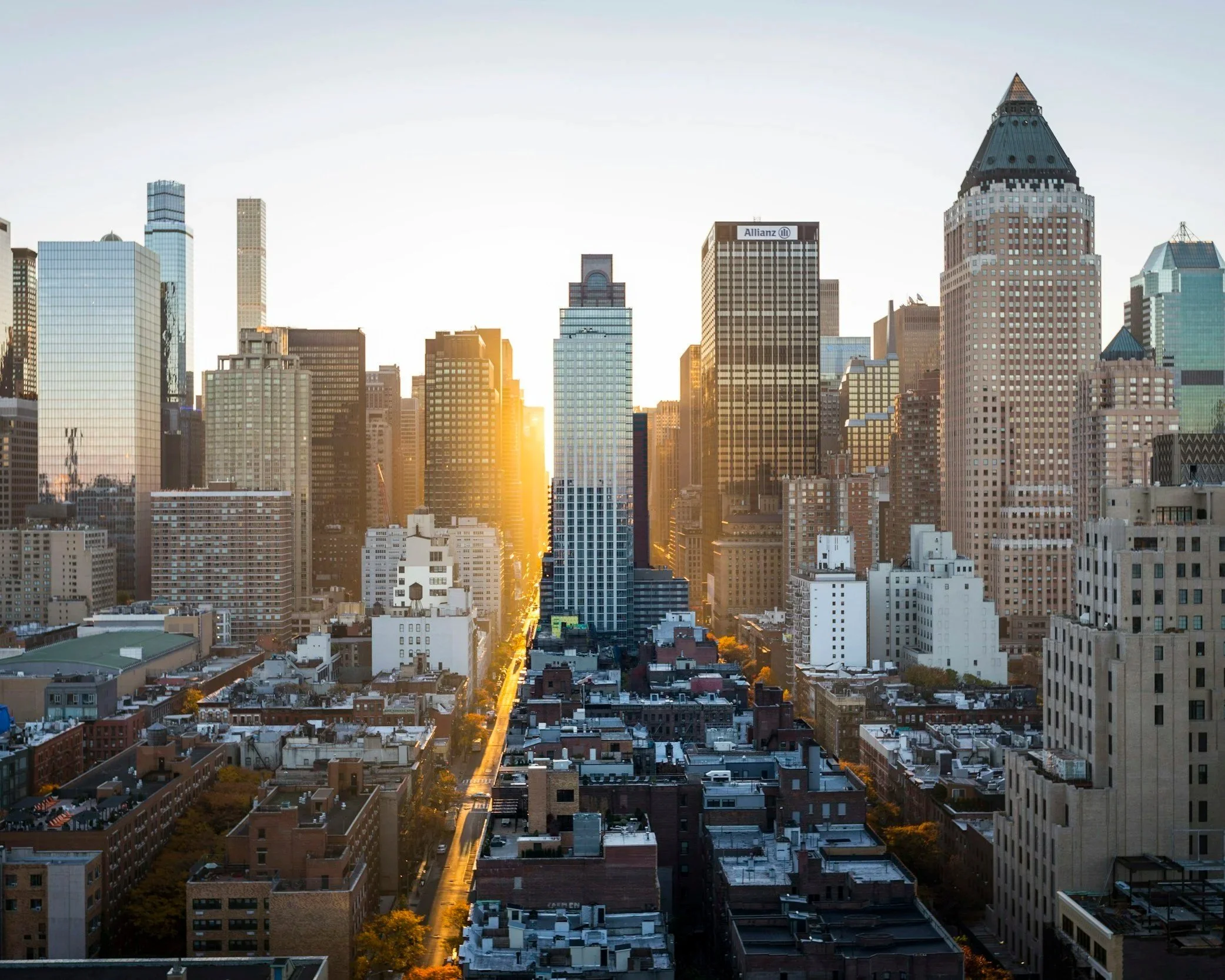 Sunset over a city skyline with tall buildings, including the Allianz building, in downtown. The sky is clear with the sun setting, casting a warm glow on the buildings.