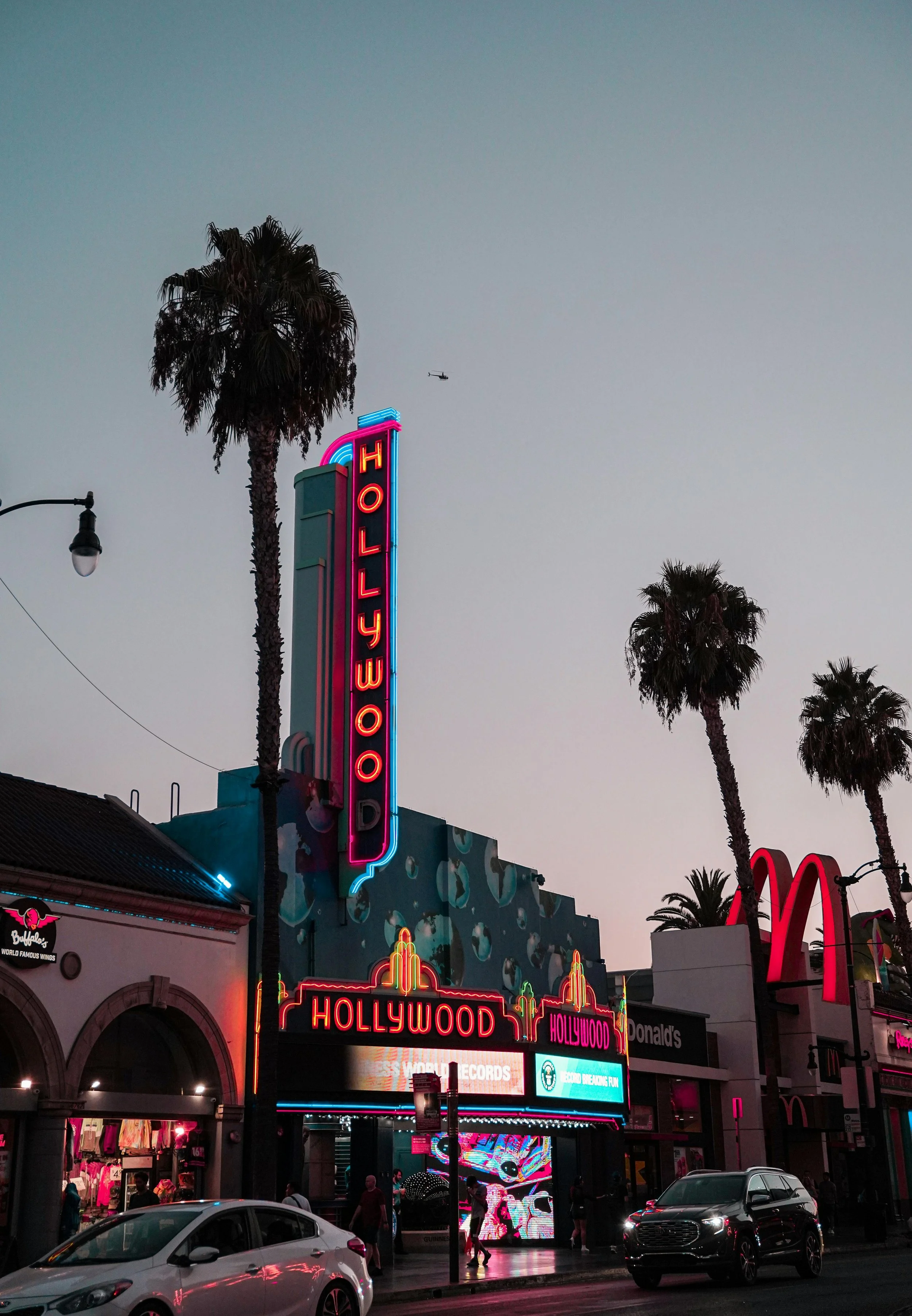 Nighttime scene of Hollywood Boulevard with neon signs, palm trees, and people walking. Visible businesses include a McDonald's and a record store, with cars on the street and a helicopter in the sky.