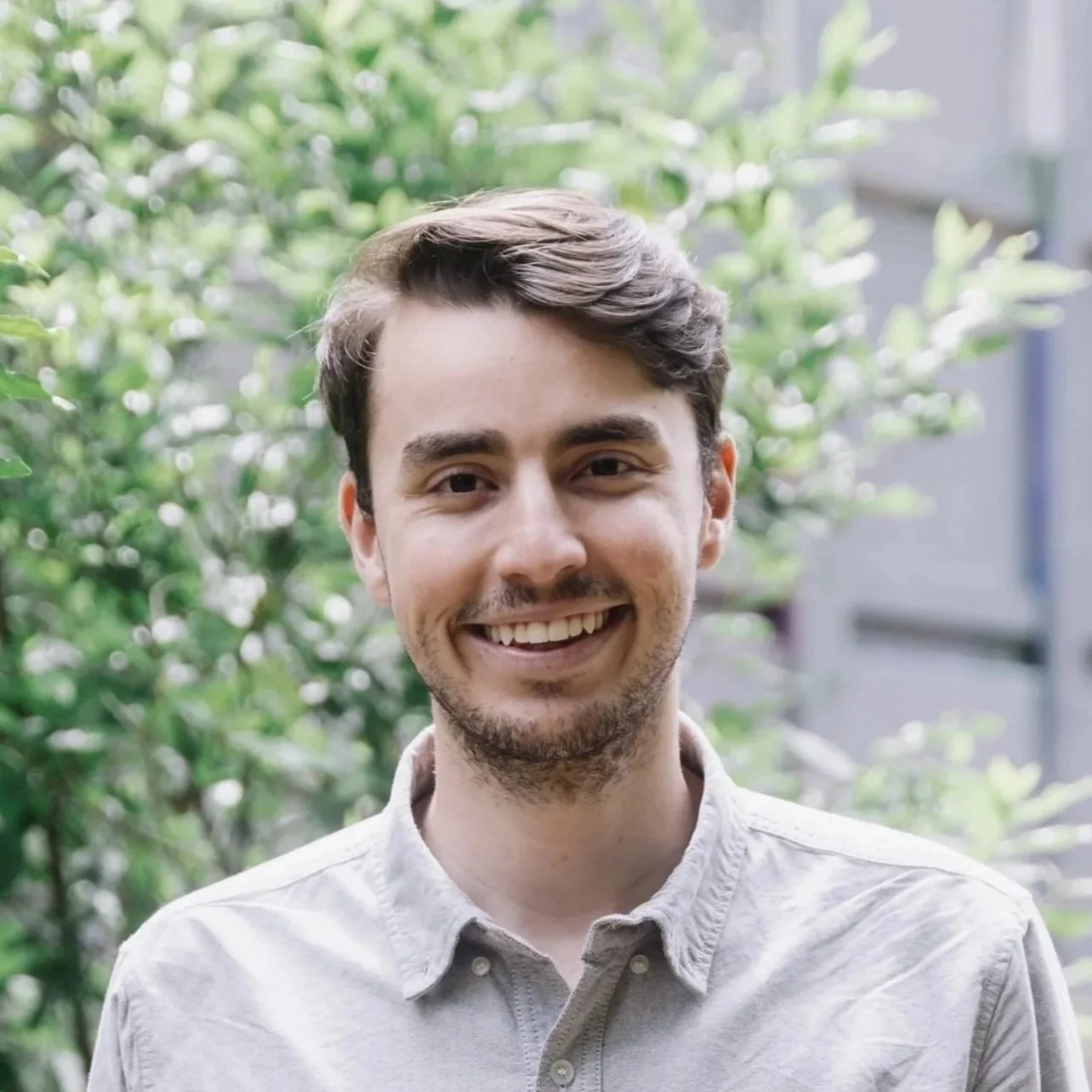 Photo of Agustín Soto, a Colombian young man with wide smile, brunette hair and a short beard standing in front of some blurred olive trees.