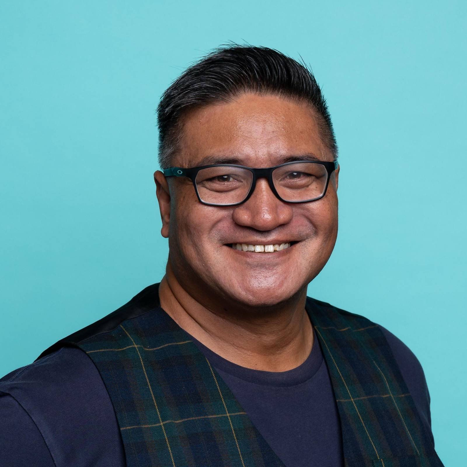 Photo of Sei Brown, a Pasifika man with a wide smile, black hair and black glasses standing in front of a bright blue background. 