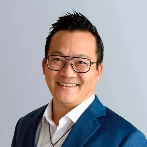 Photo of Alec Tang, a man of East Asian appearance wearing a blue suit and white shirt, with black glasses standing on front of a grey background.