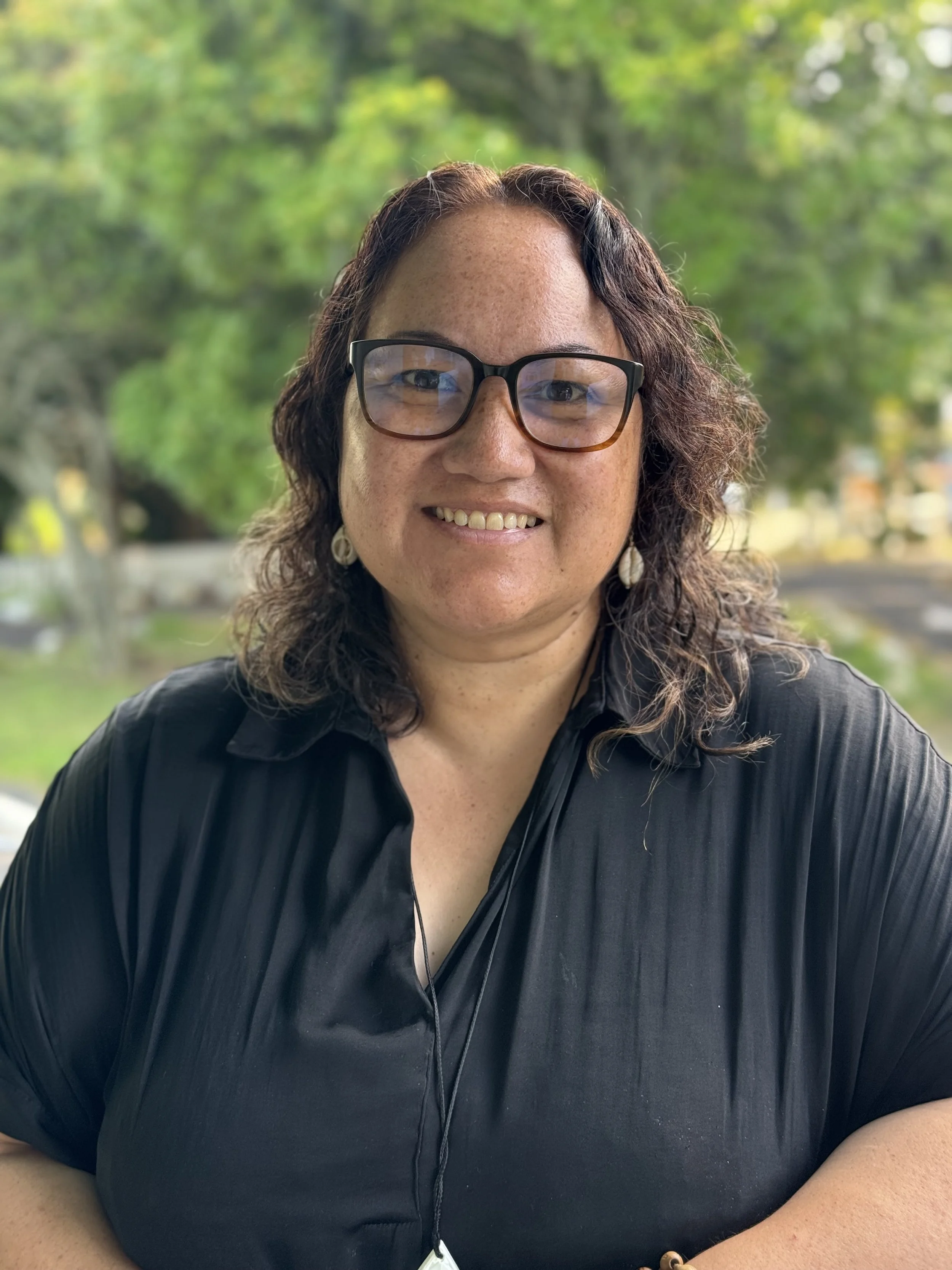 Headshot of Cecelia Vakameilalo-Kioa, a Tongan-Samoan woman wearing a black shirt and black glasses with shoulder-length wavy hair smiling at the camera.
