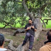 Headshot of Zane wedding, a Māori man lying on the horizontal trunk of a Pohutukawa tree gesturing to himself with his hands as he speaks.