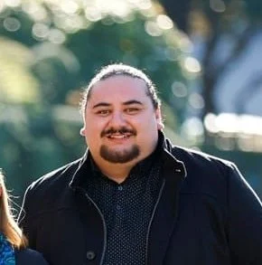 Photo of Fred Wilson, a Māori man with black hair pulled back into a pony tail and a moustach and goatee, wearing a black jacket standing in front of a blurred background of trees.