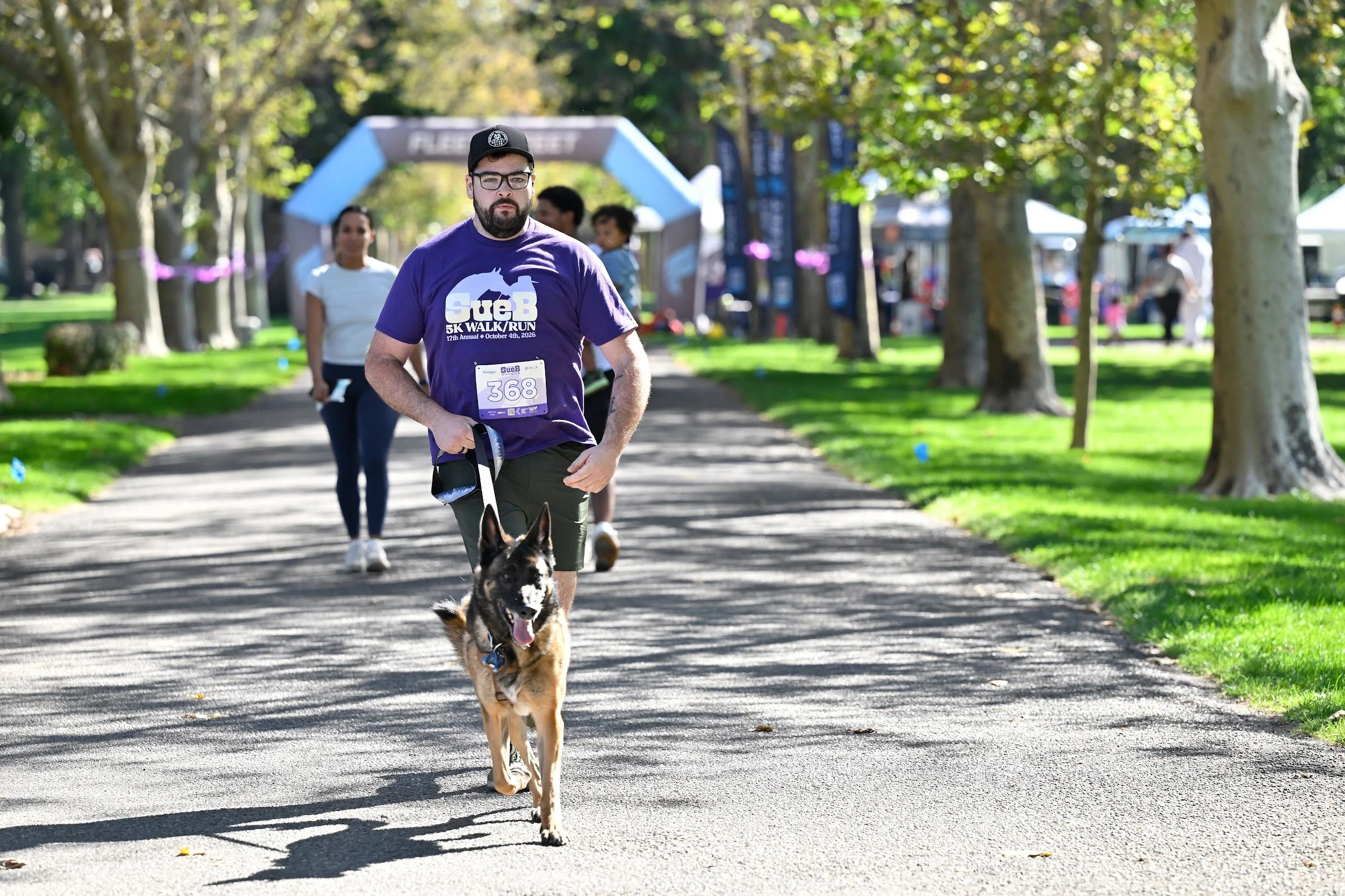 A SueB runner in a purple shirt running with a dog toward the finish arch