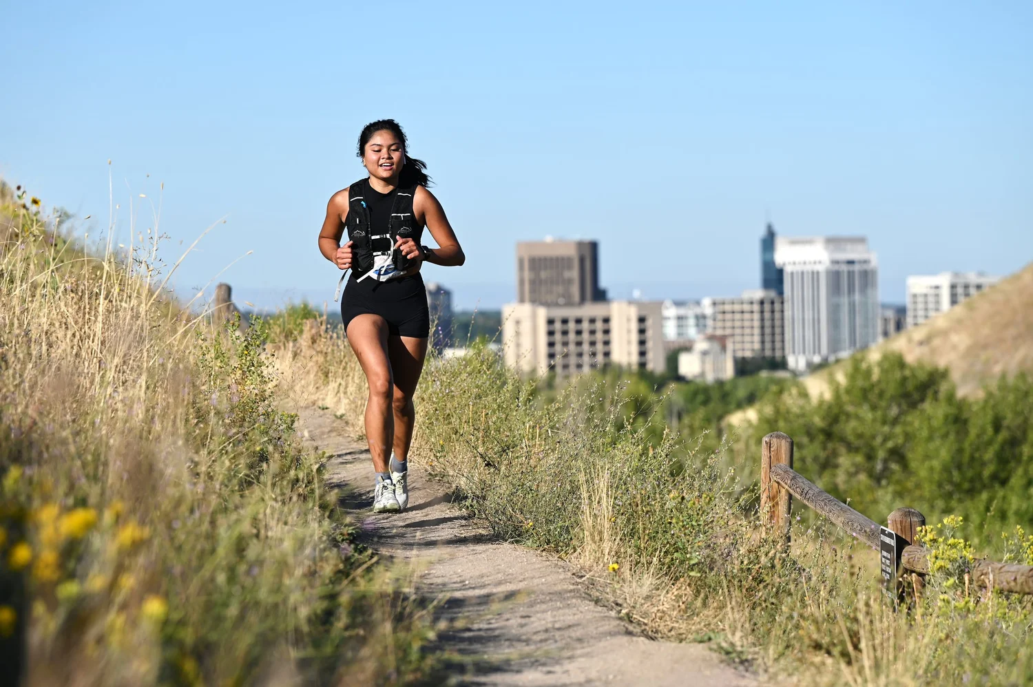 A trail runner smiles on a foothills singletrack with the downtown Boise skyline behind her