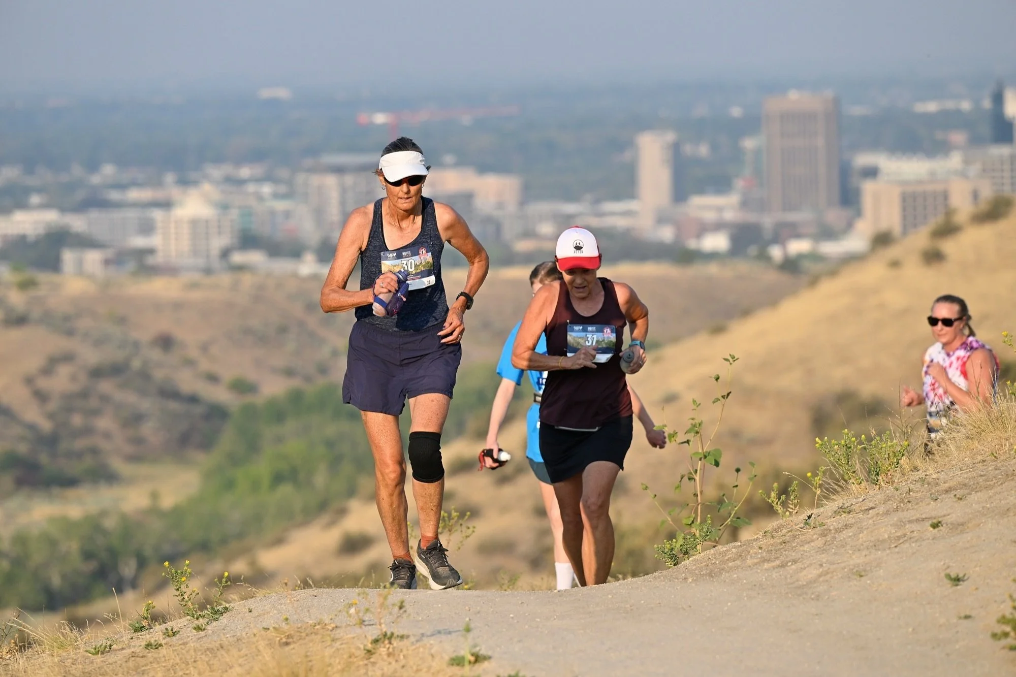 Two runners climb a Military Reserve trail with downtown Boise behind them at the Foothills XC12K
