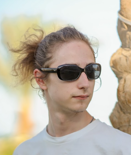 Young man with curly hair wearing sunglasses and a white shirt outdoors on a sunny day.
