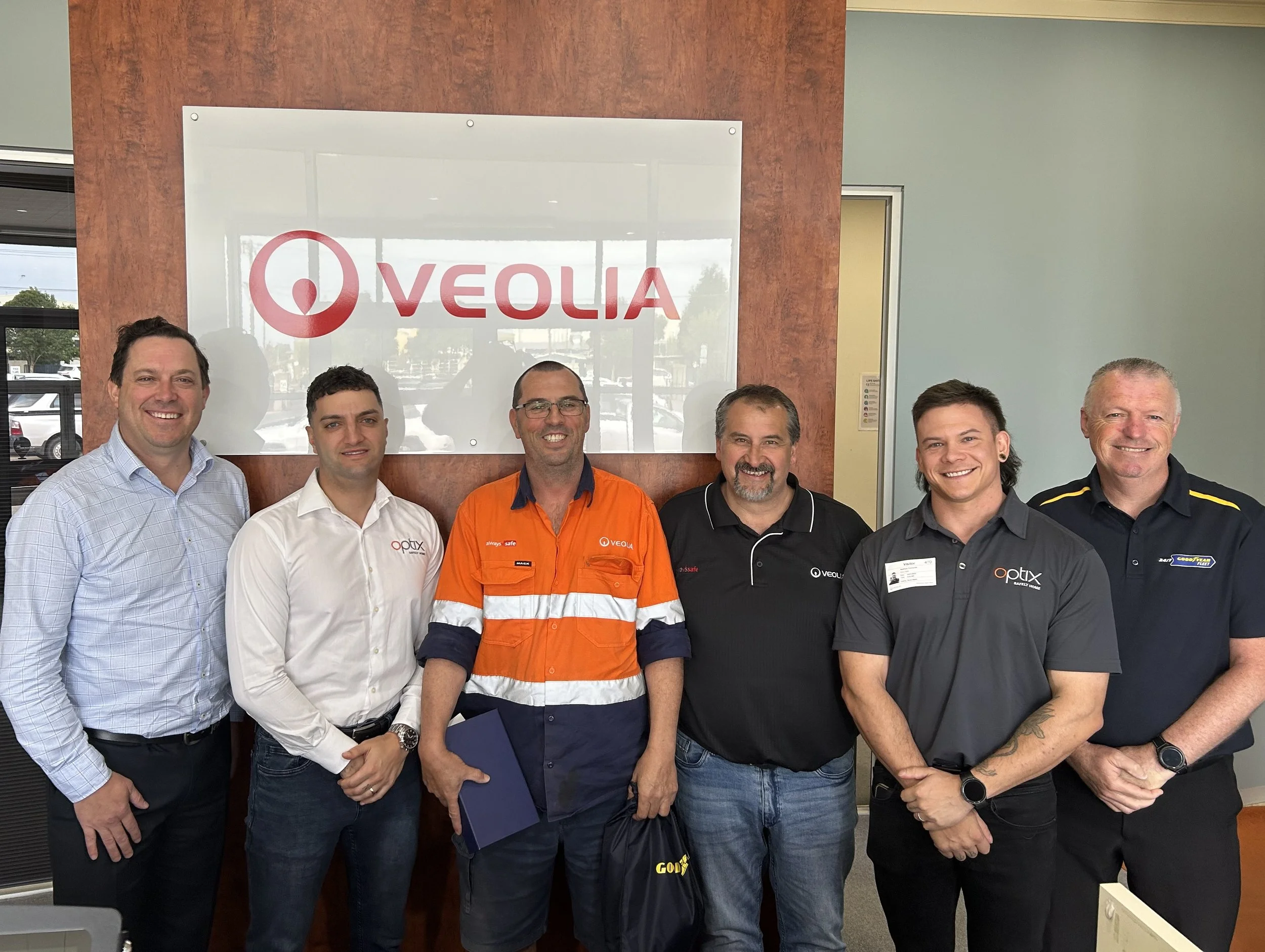 Group of six men standing in front of a Veolia sign, wearing various branded shirts and smiling at the camera.
