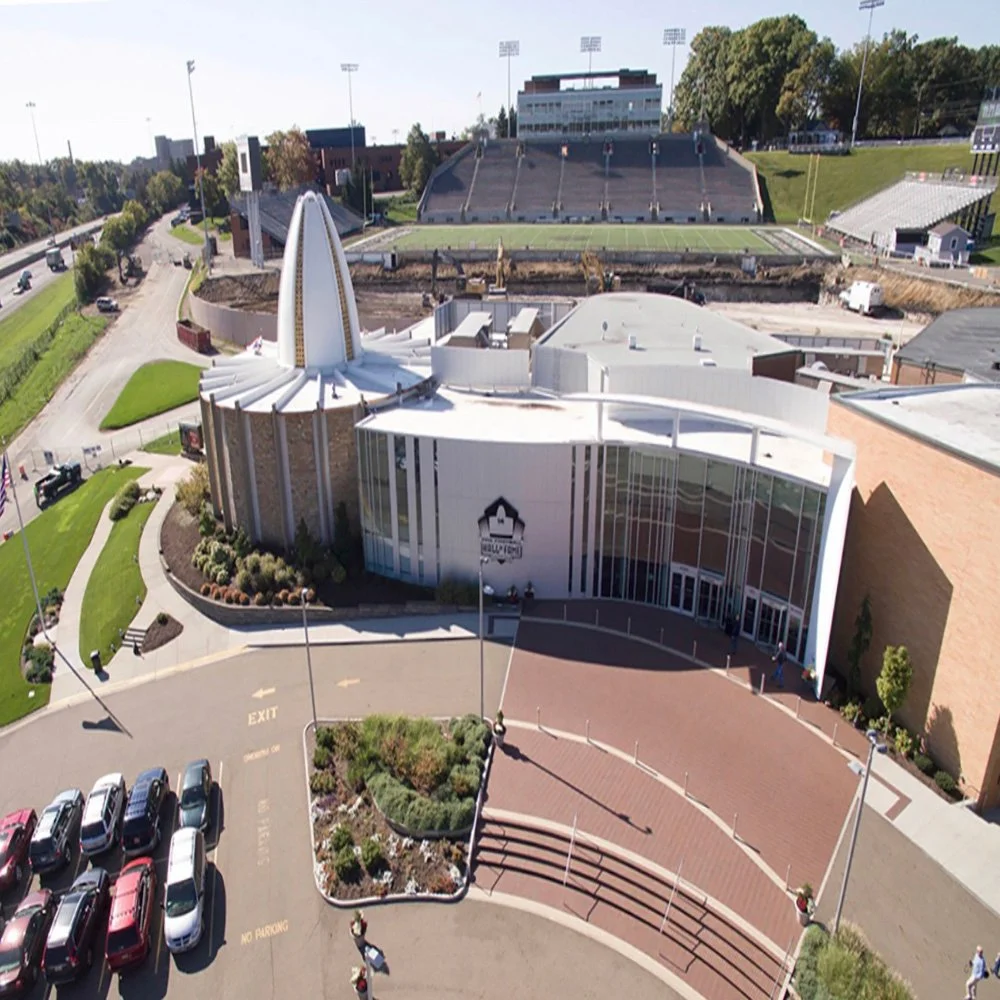 Aerial view of a stadium and a modern building with a church-like steeple, parking lot, and landscaped area in a city setting.