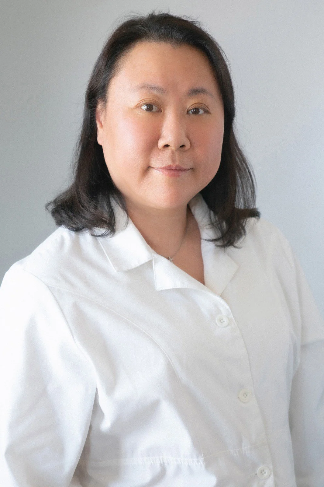 Portrait of a woman with shoulder-length dark hair, wearing a white button-up shirt, against a plain light gray background.