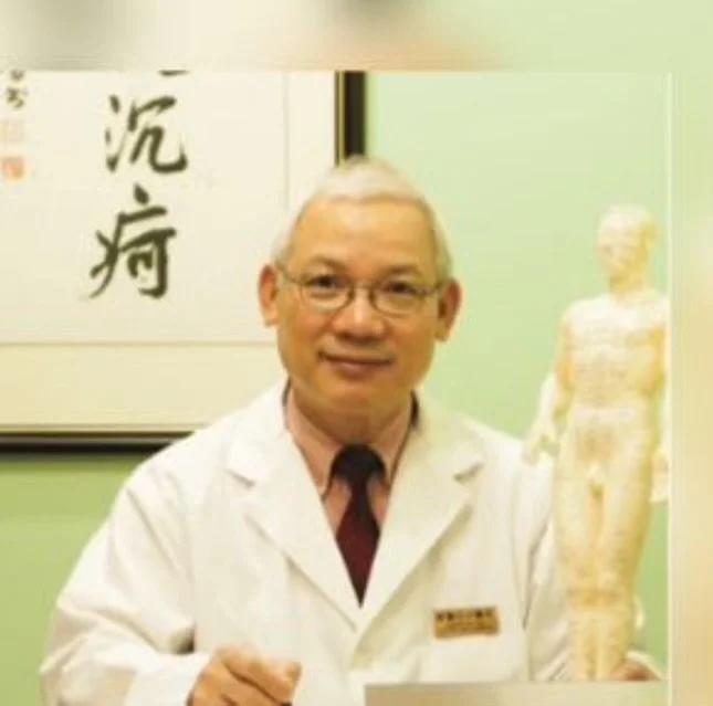 A doctor with glasses and gray hair holding a model of a human body, standing in front of a green wall with Asian calligraphy.