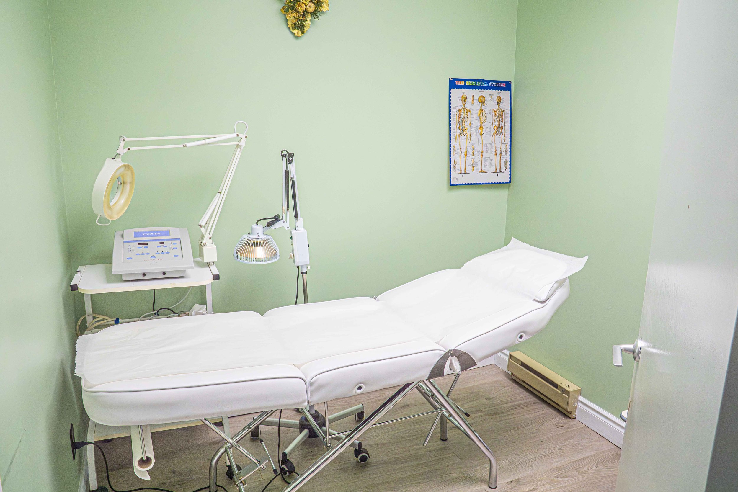 Empty medical examination room with a padded examination table, medical equipment, a poster of the skeletal system on the green wall, and a wooden floor.