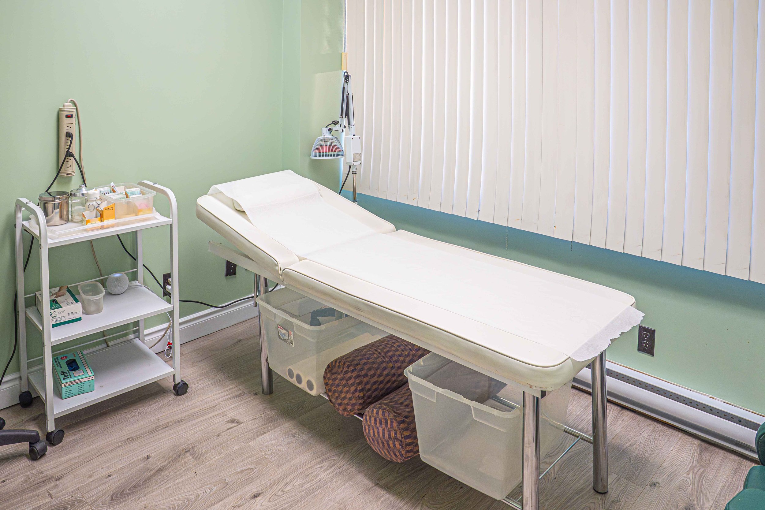 Empty hospital examination room with a bed, medical supplies on a cart, and closed vertical blinds.