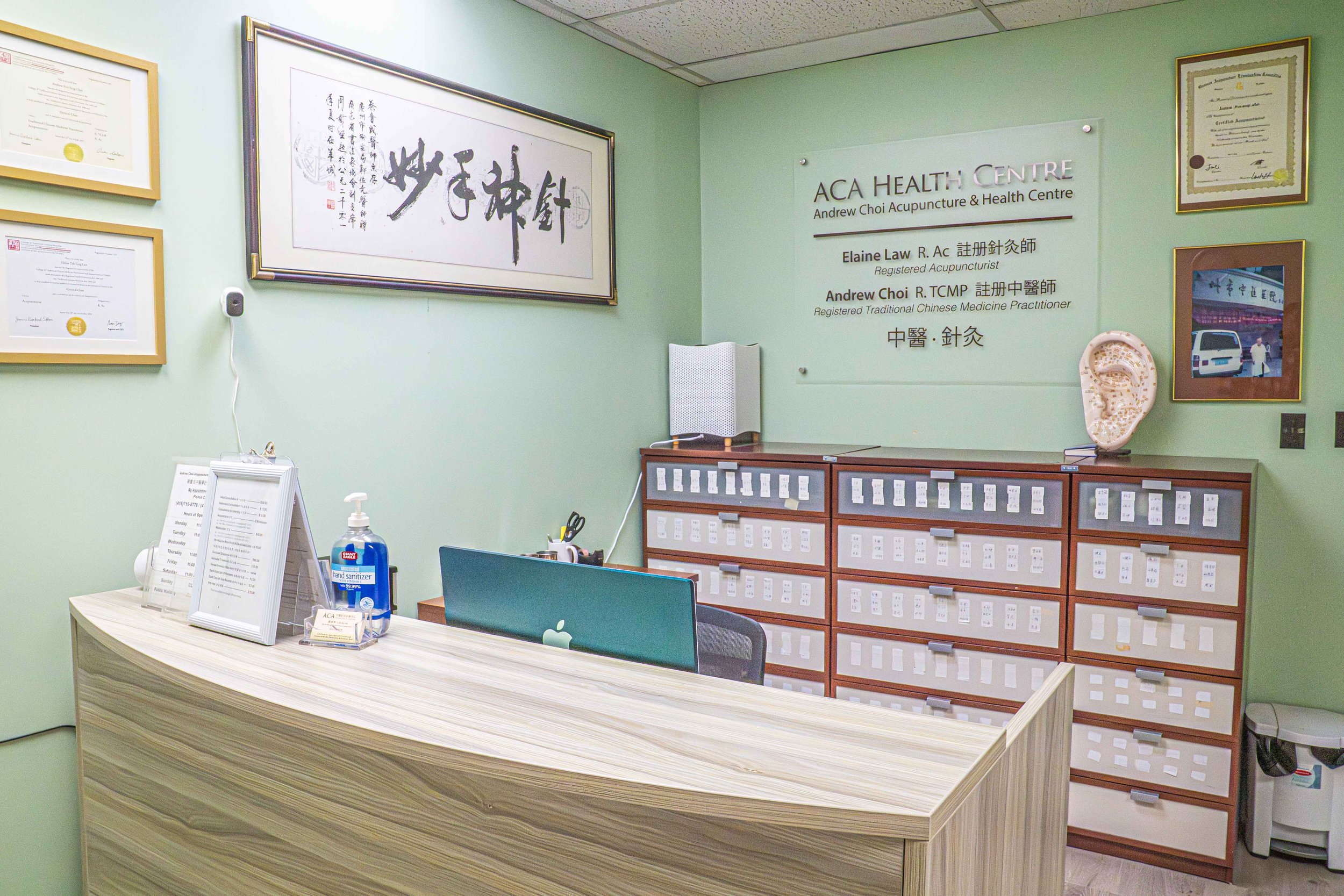 Reception area of a health center with a wooden desk, a computer, hand sanitizer, and framed certificates and artwork on a light green wall.
