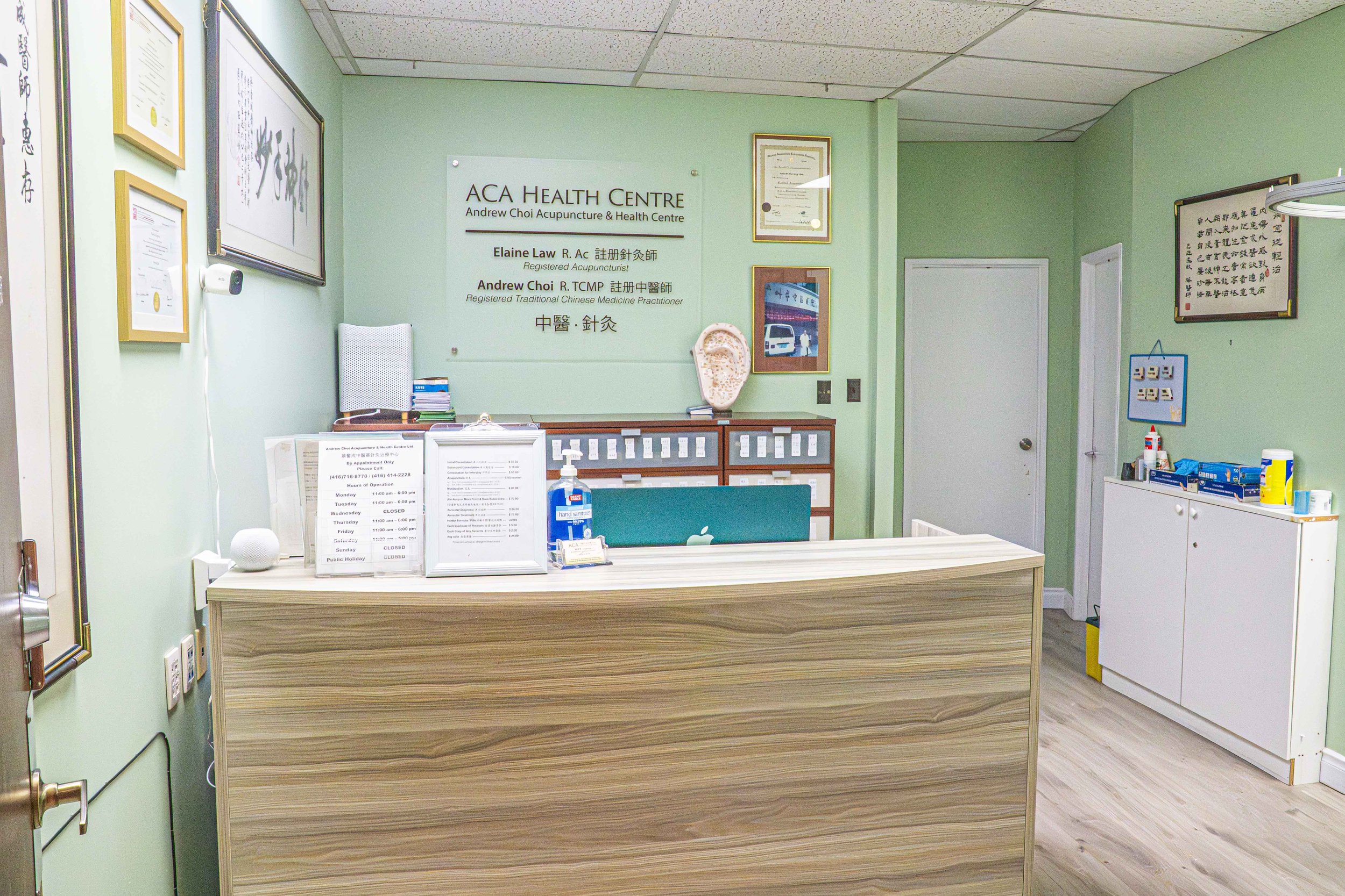 Reception area of a healthcare center with a wooden desk, a computer, hand sanitizer, certificates on the wall, and a sign reading 'ACA Health Centre' behind the desk.