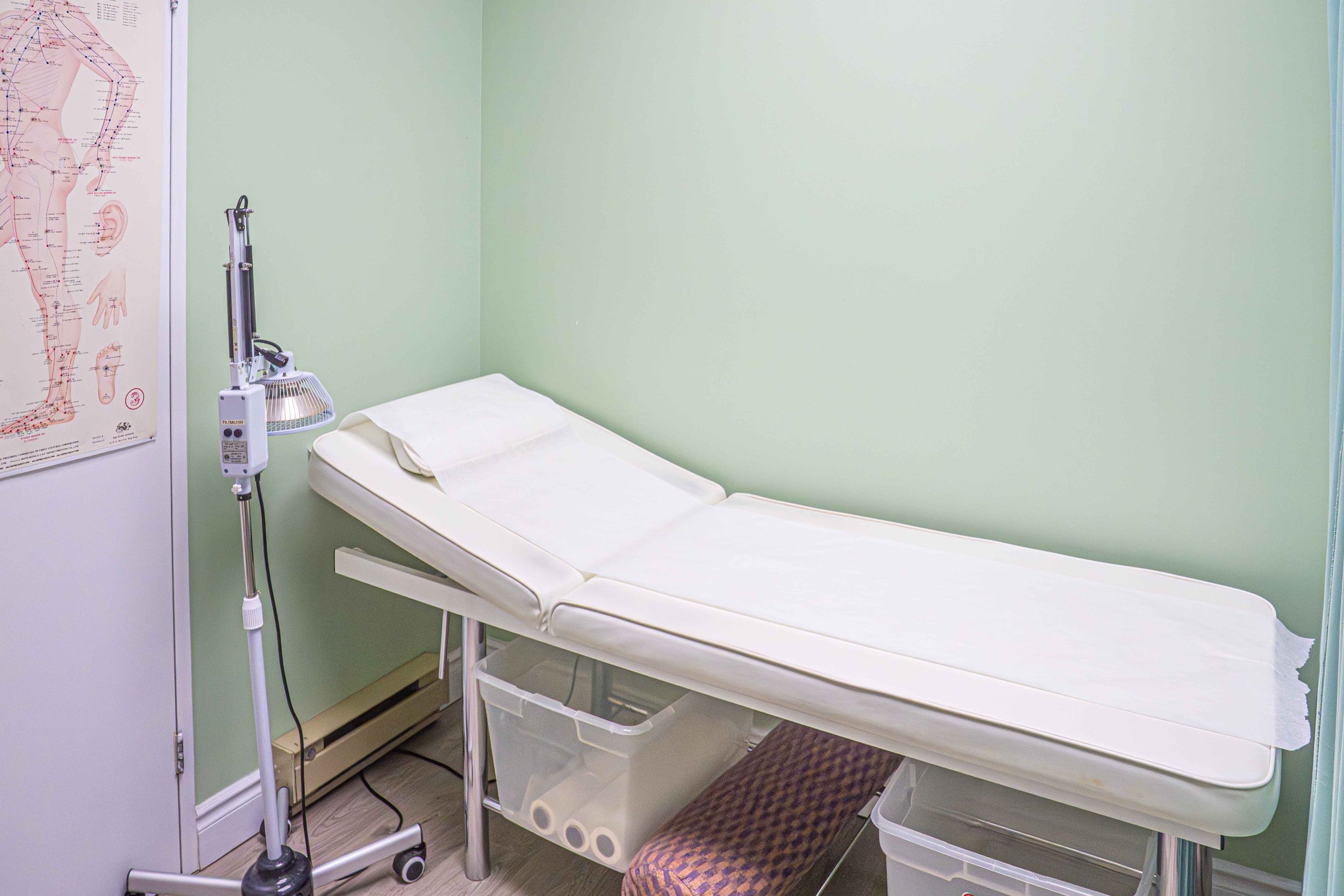 An empty examination room with a medical bed covered in a white sheet, medical equipment, and a poster of acupuncture points on the wall.