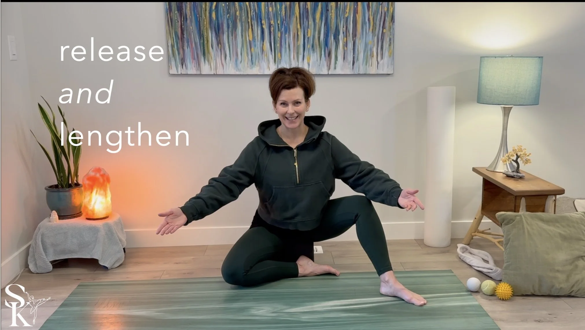 A woman practicing yoga indoors, in a squat position with arms extended outward. She is smiling and wearing a black hoodie and black leggings. The room is decorated with a colorful abstract painting, a blue lampshade, a salt lamp, and a small table with a plant. There are yoga props on the floor, including a ball and textured massage balls.
