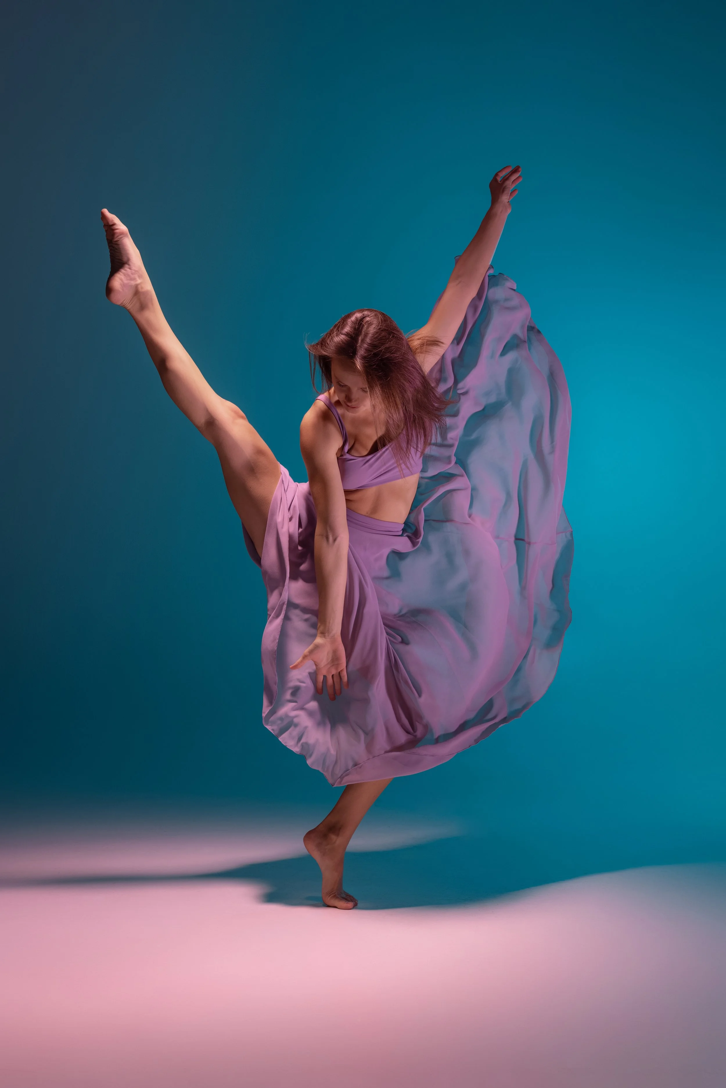A woman performing a dance pose with one leg raised high, wearing a flowing lavender dress, against a gradient background of blue and pink.