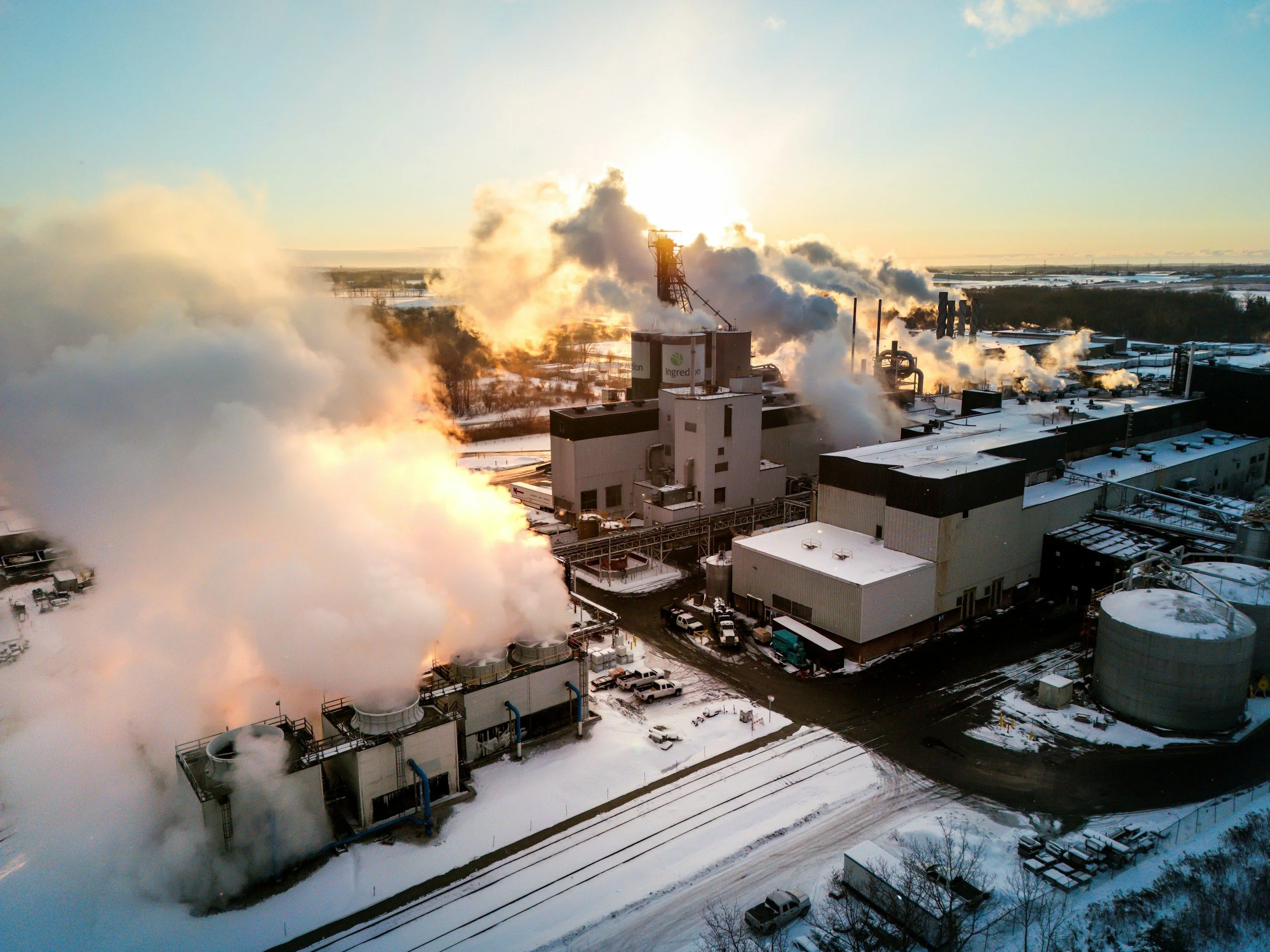 An industrial plant with multiple smokestacks emitting steam and smoke, situated in a cold, snowy landscape during sunset.