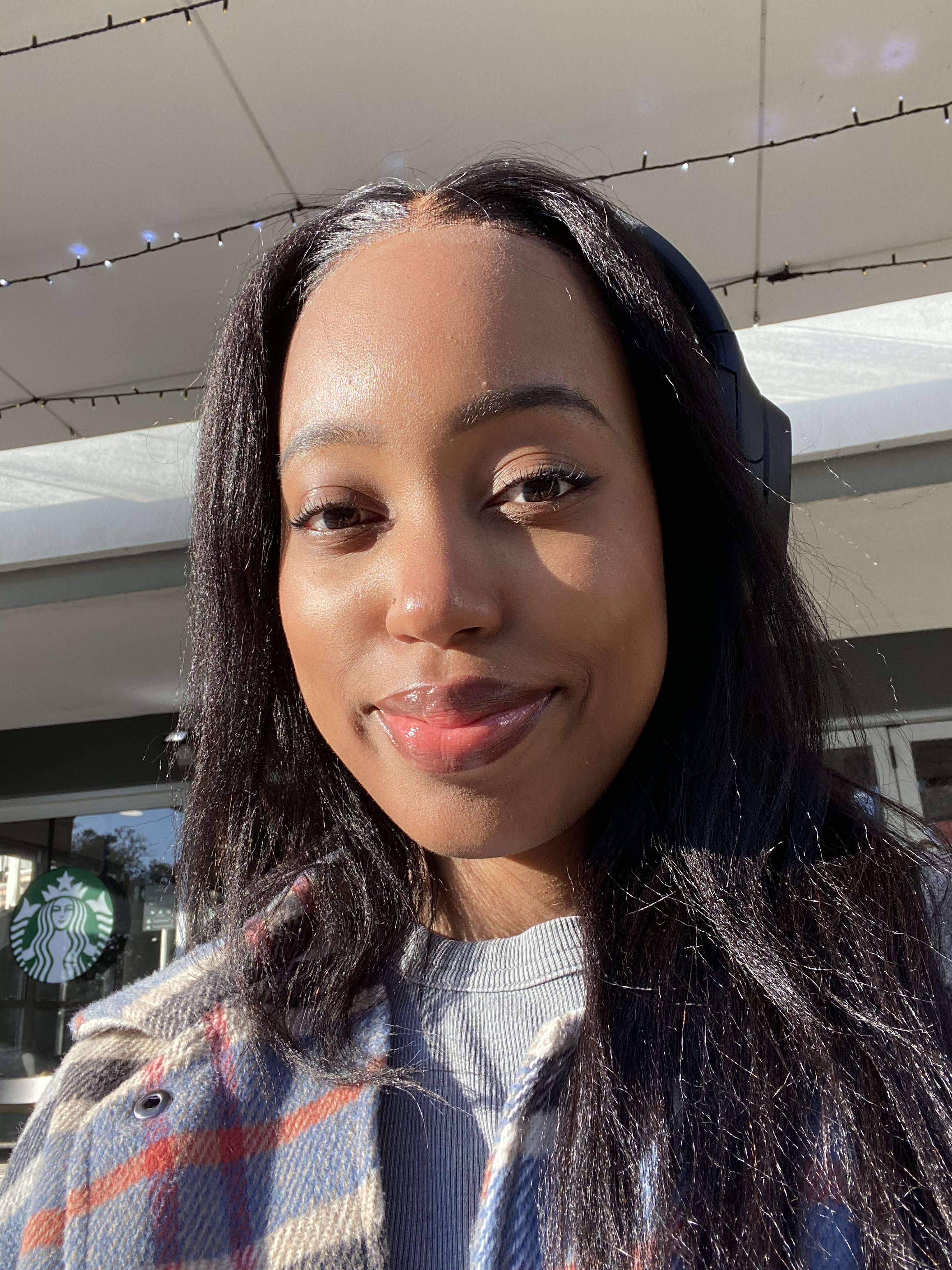 Young woman with dark hair wearing headphones, smiling outdoors in front of a Starbucks store, sunlit.