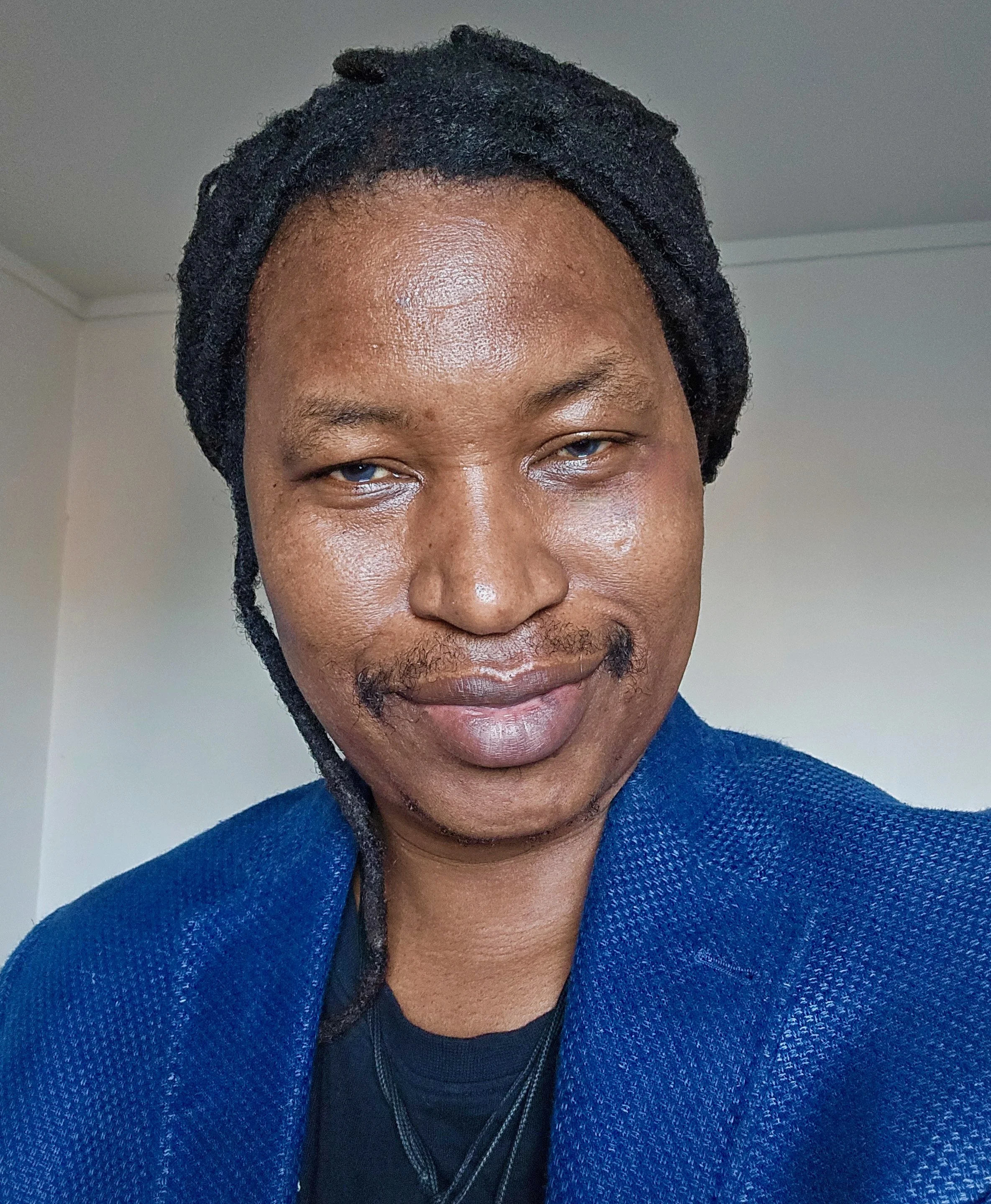 Close-up portrait of a man with dreadlocks, wearing a blue jacket, slightly smiling, against a plain background.