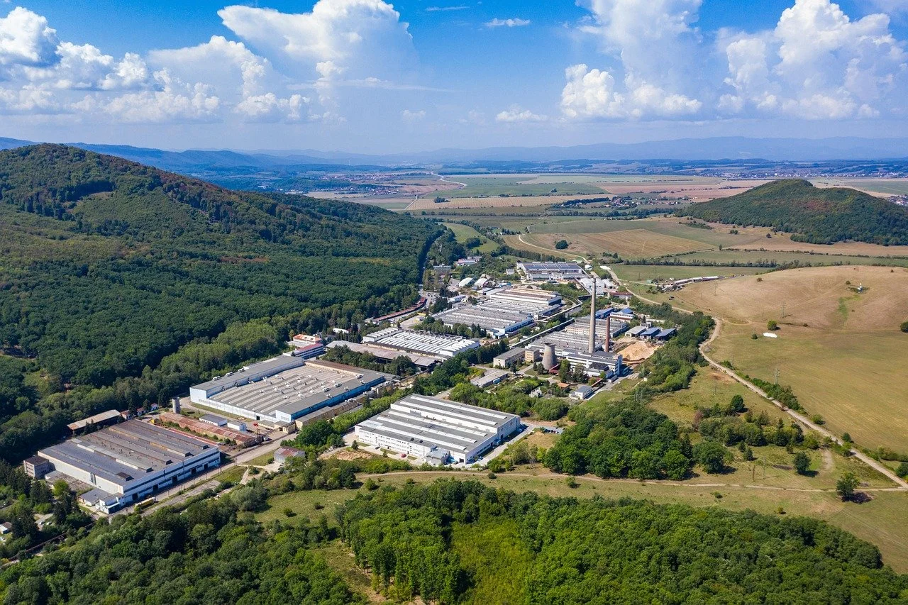 Aerial view of an industrial complex surrounded by green forests and rolling hills under a partly cloudy sky.
