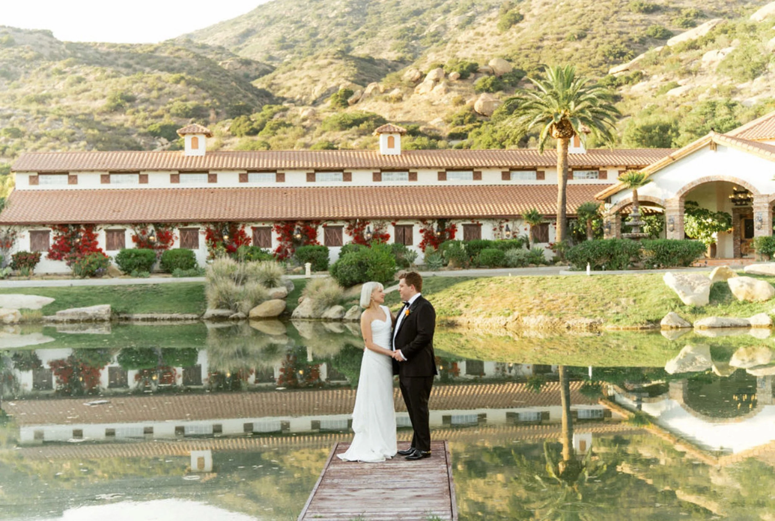 A bride and groom standing on a small dock by a reflective pond, with a large house, palm trees, and hills in the background.