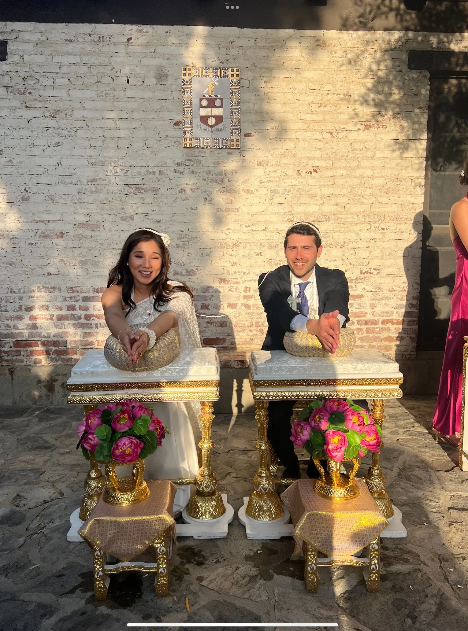 A smiling woman and man dressed in traditional clothing, sitting behind decorated tables with gold ornaments and pink flower arrangements, in front of a brick wall with a crest or emblem on it, during a celebration or ceremony.