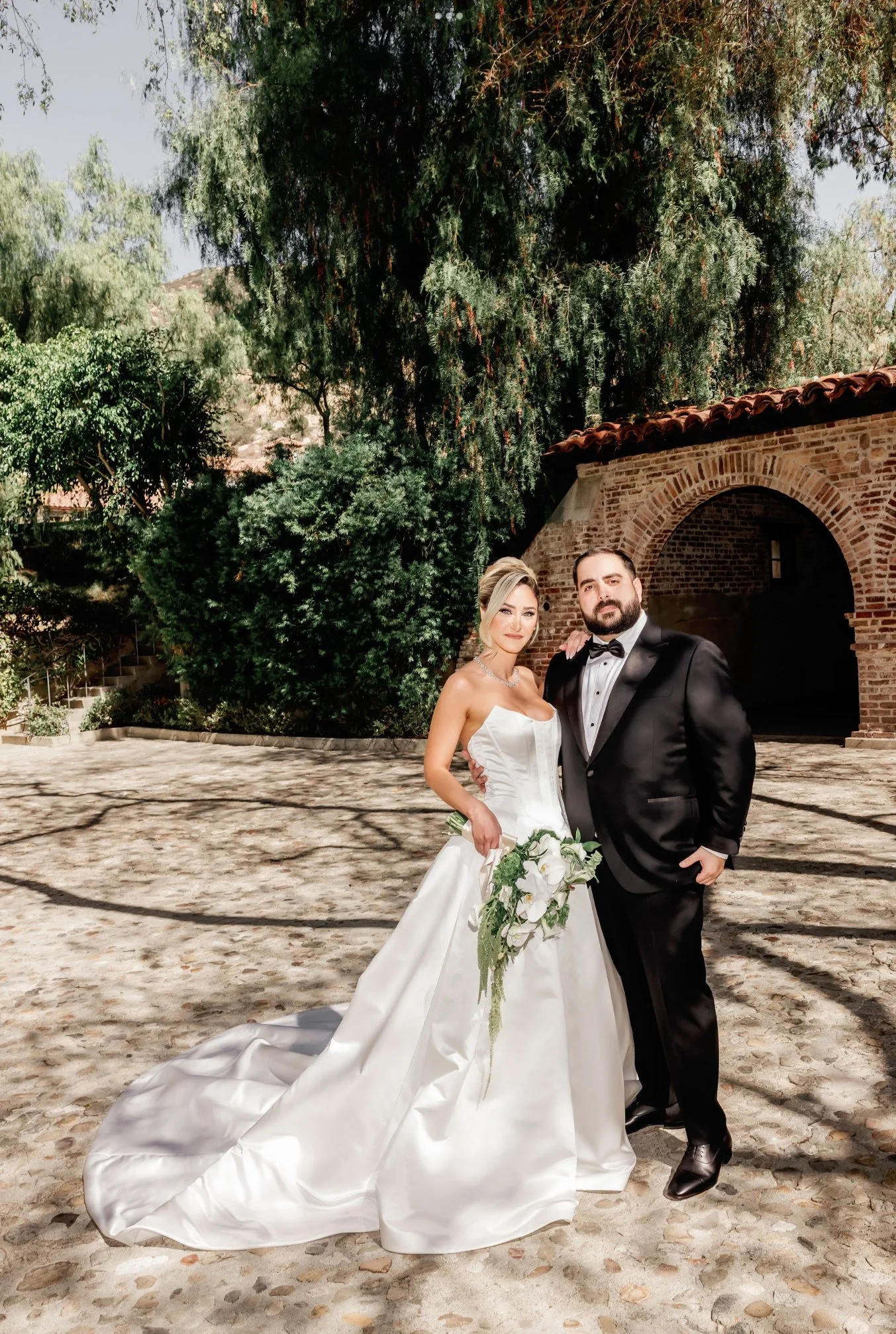 A bride and groom in wedding attire posing outdoors on a cobblestone pathway with trees and a brick archway in the background.