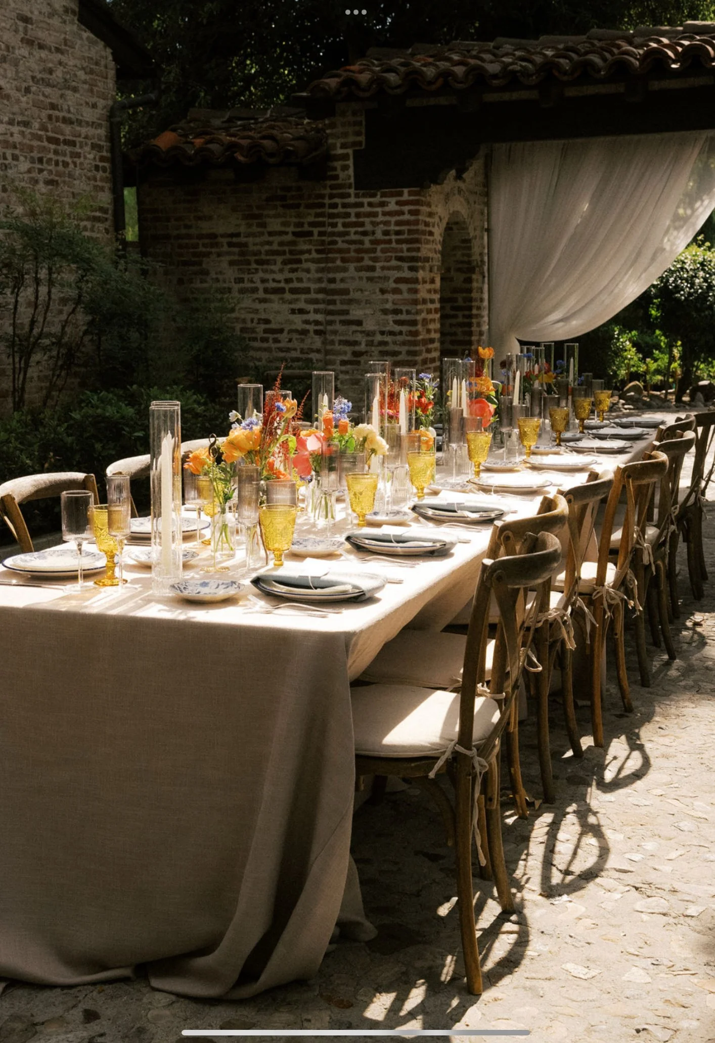 A long outdoor table set for a meal with floral centerpieces, yellow-tinted glassware, and white plates, located on a stone patio next to a rustic brick and stone building with a tiled roof and draped fabric decoration.