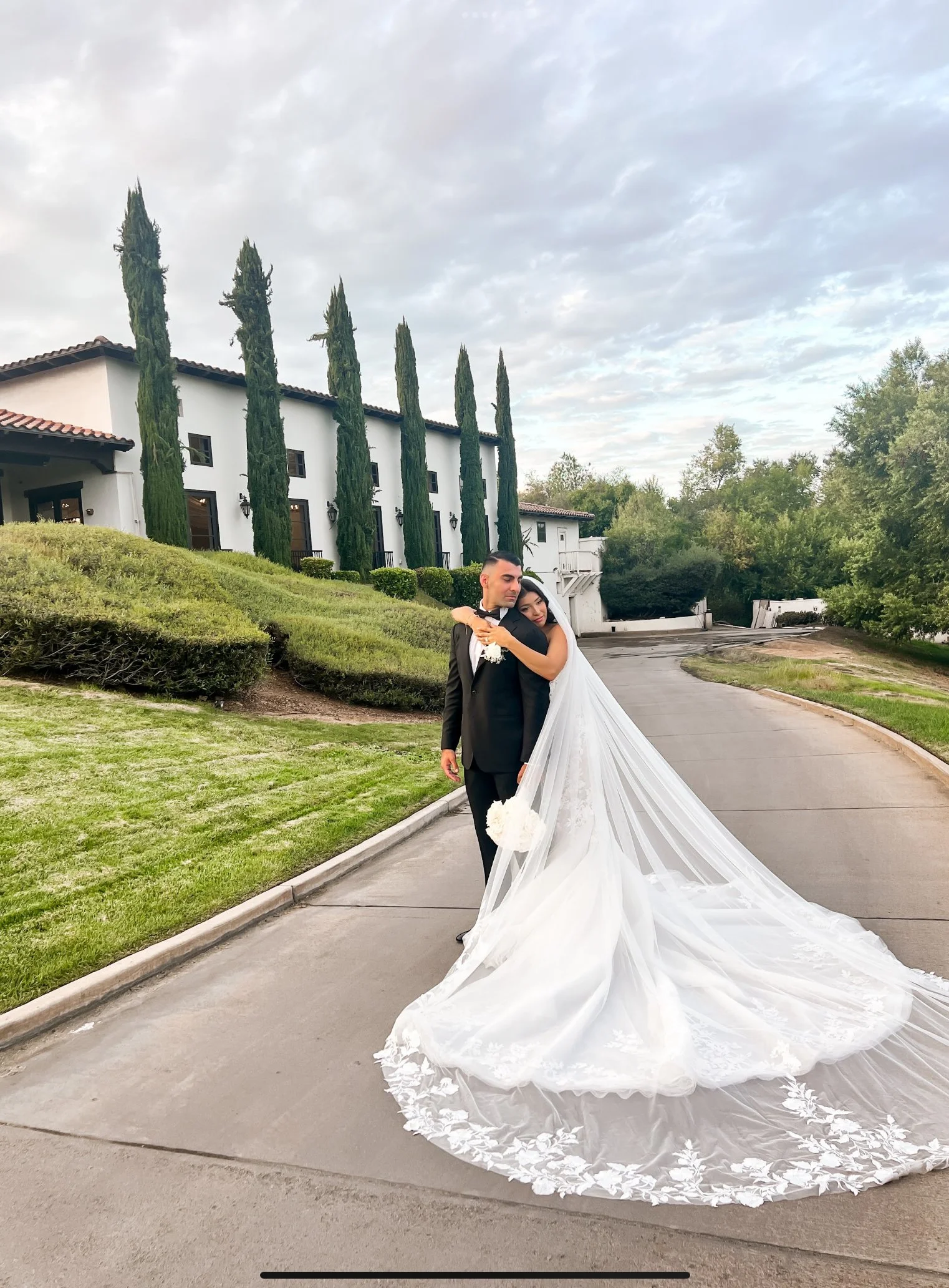 A bride and groom standing outside on a curved driveway in front of a white building with tall cypress trees. The bride is in a white wedding gown with a long train, and the groom is in a black tuxedo. The bride is hugging the groom. The sky is partl