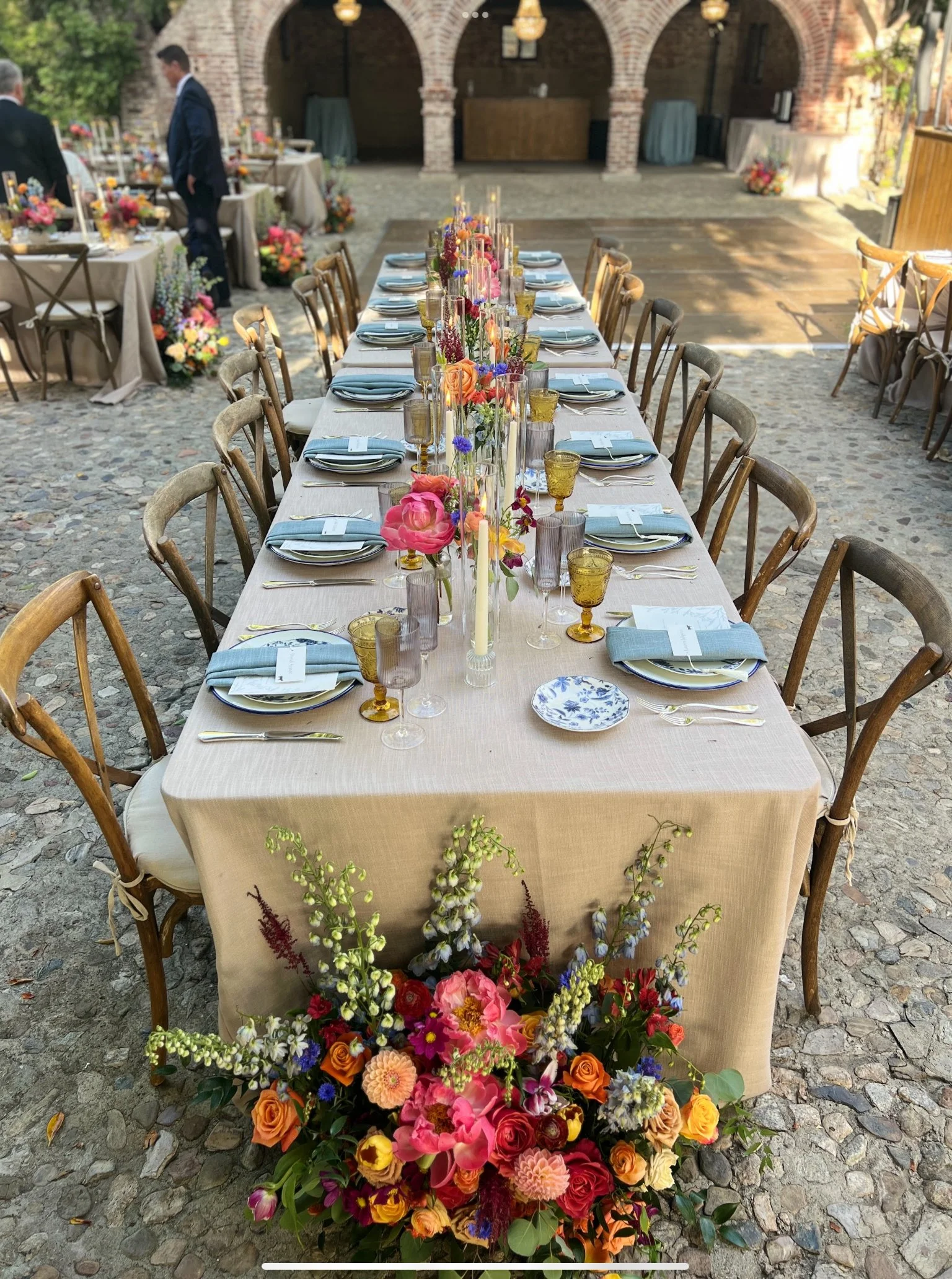 Elegant long banquet table decorated with colorful flowers, candles, and glassware on an outdoor stone-paved area near brick archways, with a floral centerpiece at the end and additional tables in the background.