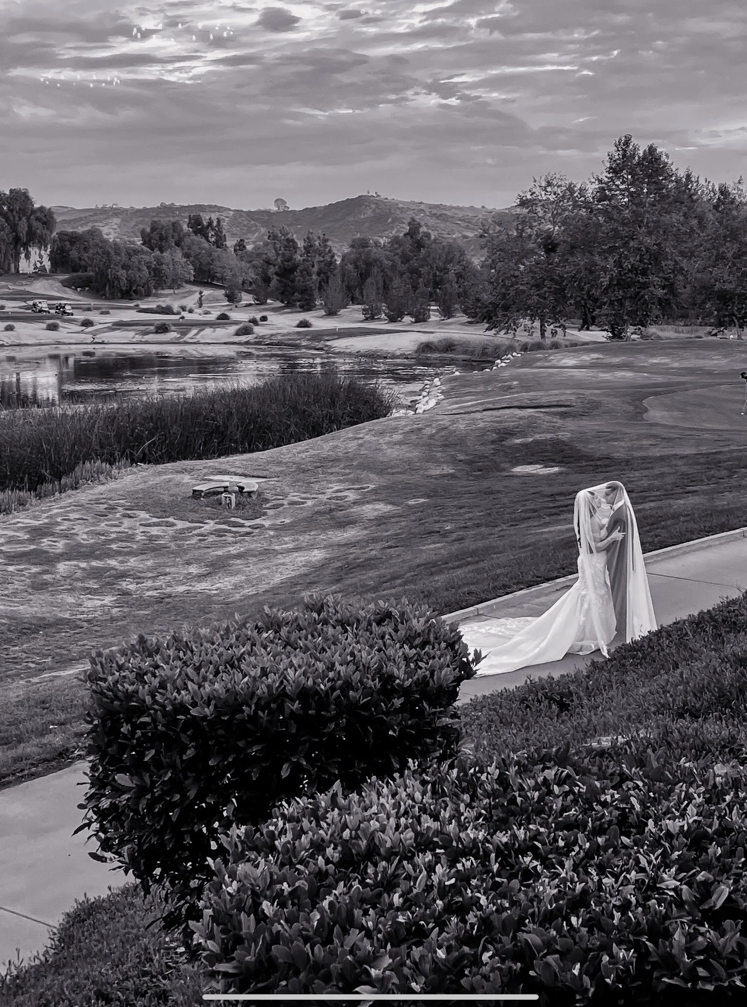 A black and white photo of a statue of a woman in a flowing dress standing on the sidewalk near a shrub. Behind her, there's a lawn, trees, a small pond, and rolling hills with a cloudy sky overhead.