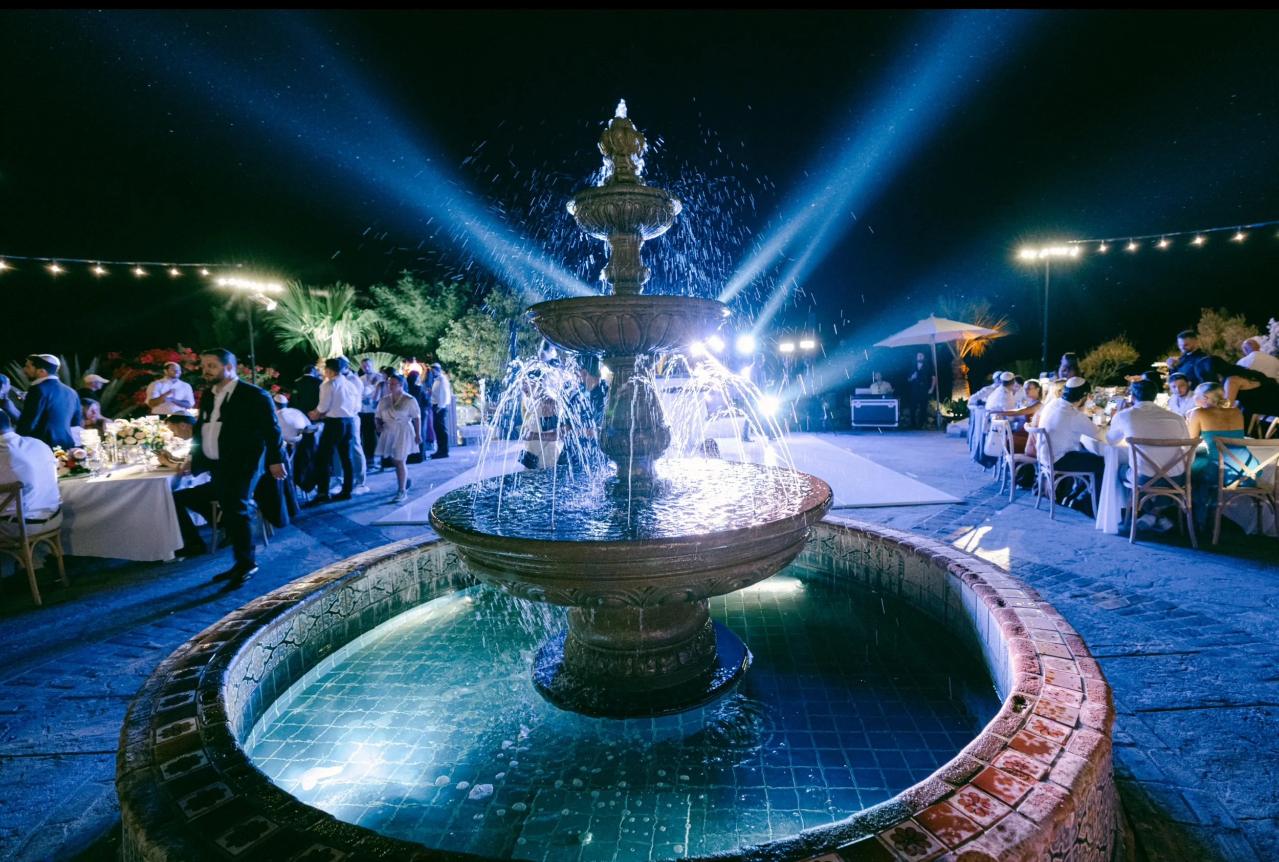 Nighttime outdoor party with a fountain illuminated by blue lighting, surrounded by tables with guests dressed in formal attire, under string lights and umbrellas.