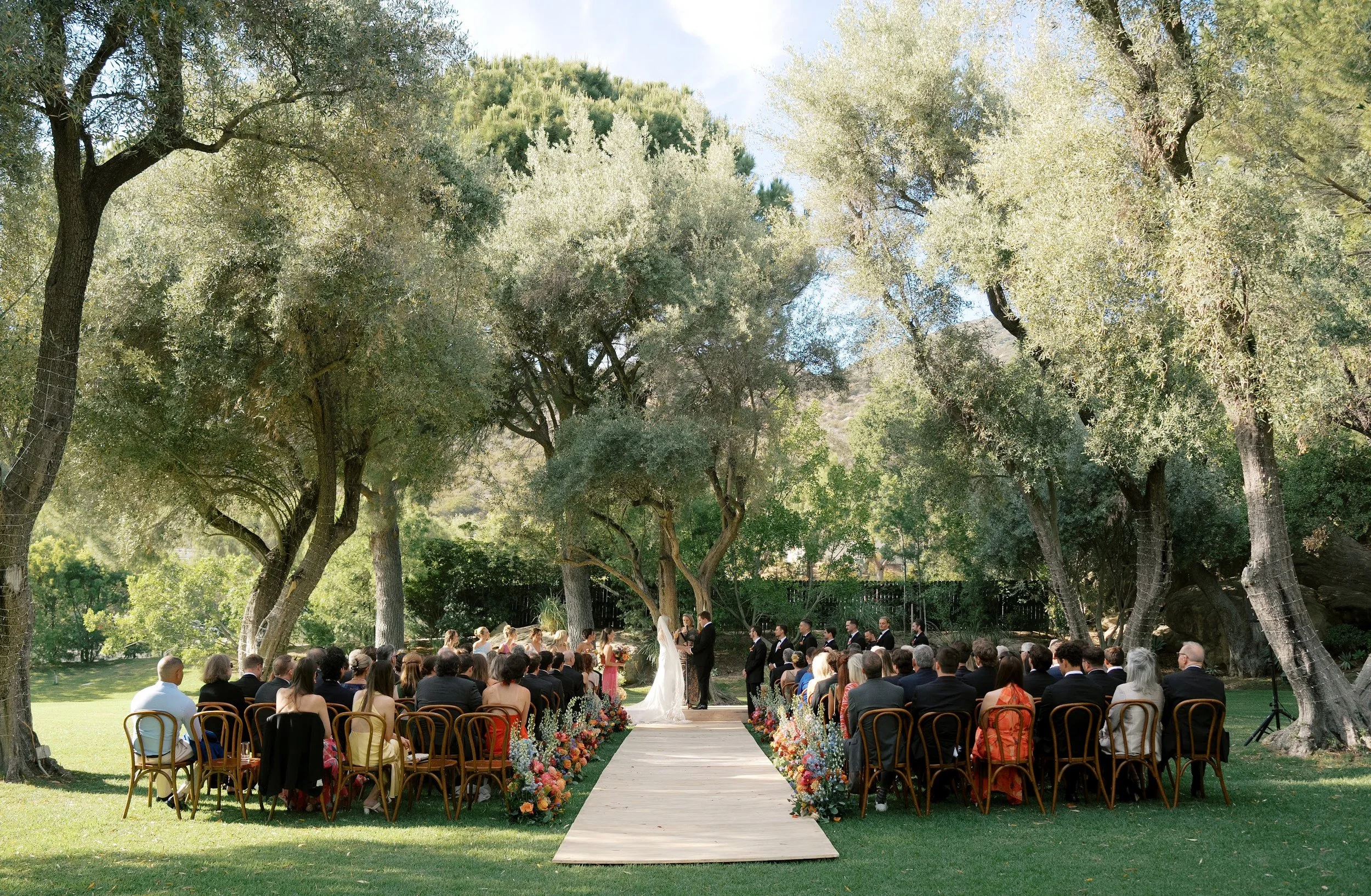 Outdoor wedding ceremony set under large trees with guests seated, a wooden aisle, and floral arrangements.