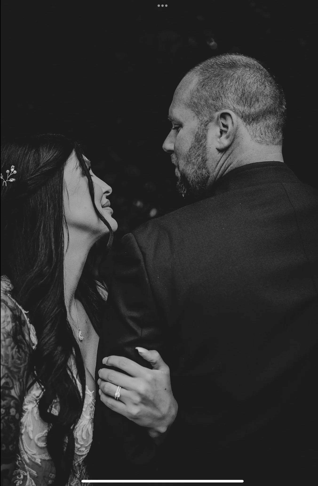 A black-and-white photo of a couple gazing into each other's eyes closely, with the woman's hand over the man's arm, showing her wedding ring and an ornate tattoo on her arm.