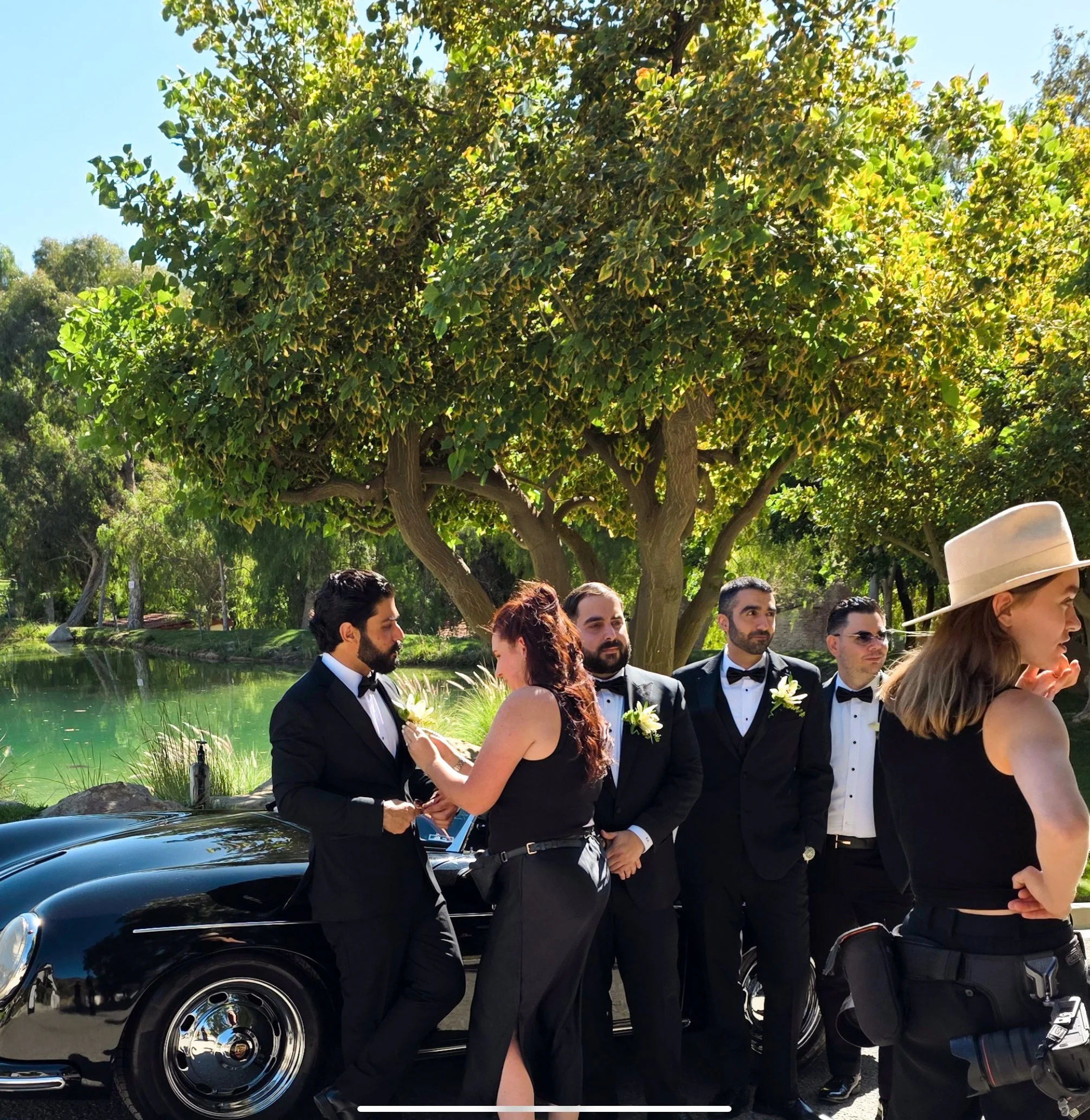 A group of people dressed in formal attire standing outdoors near a car, with a large tree and greenery in the background. It appears to be a wedding or special event.