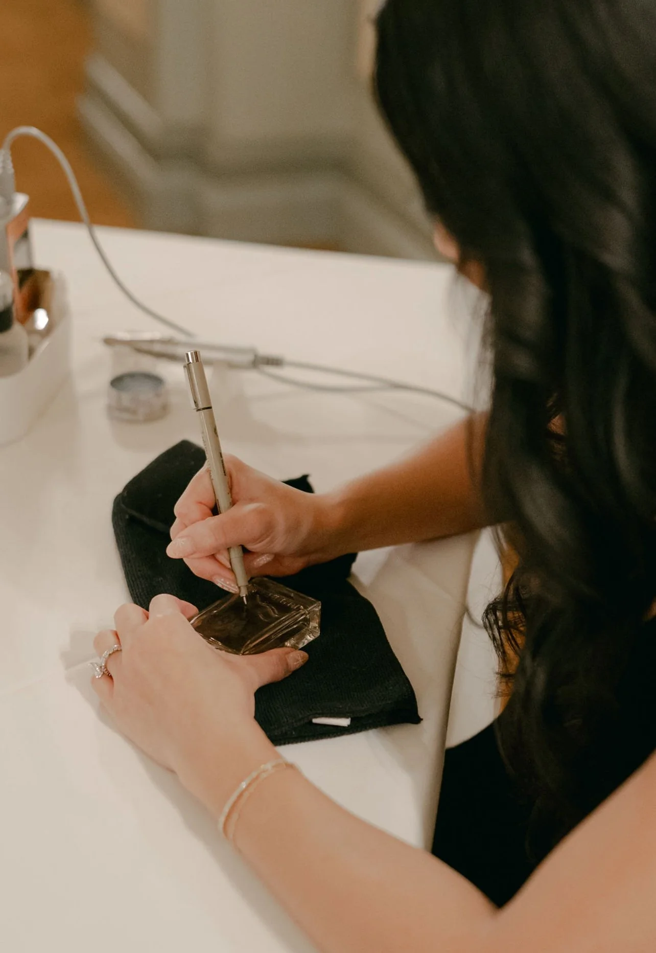 A woman with long, dark hair is sitting at a white table, writing or signing on a small, clear box with a silver pen. She is wearing jewelry, including a bracelet and rings, and has a black bag or pouch in front of her. The background shows a blurred