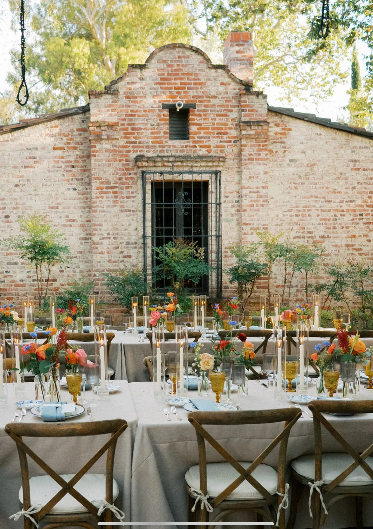 Outdoor dining setup with long tables covered in white tablecloths, decorated with colorful flowers in small vases, tall candles, and set with plates, glasses, and napkins. The background features a brick building with a small window, surrounded by g