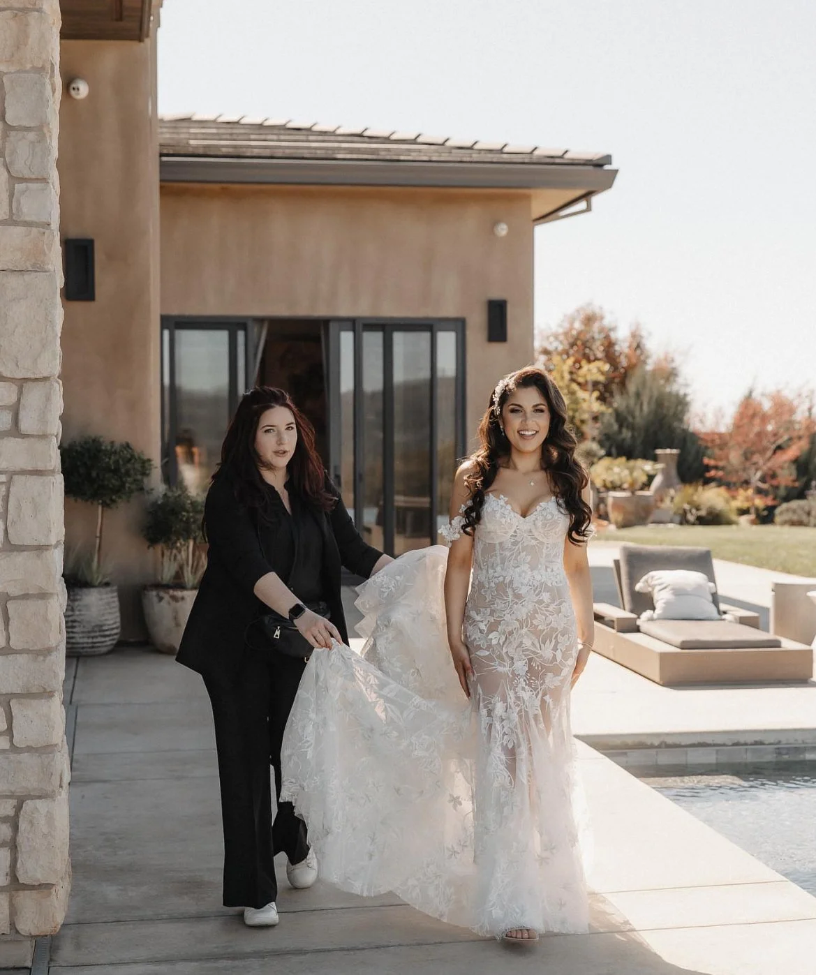 A bride in a lace wedding gown is being assisted with her dress by a woman in black outside a modern house, with seating and plants visible on the patio and trees in the background.
